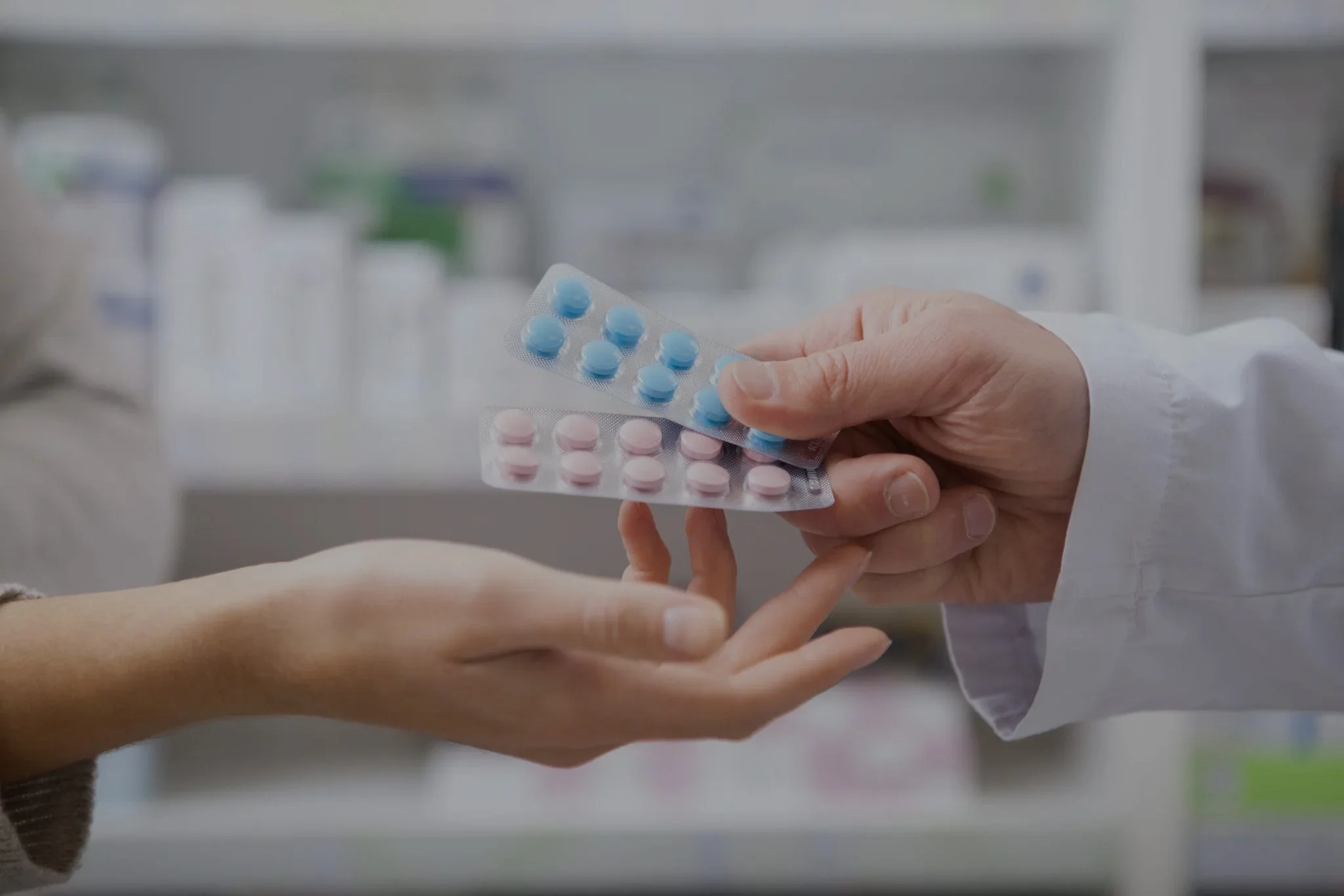A pharmacist handing pills to a customer in a pharmacy, with blister packs containing blue and pink pills.