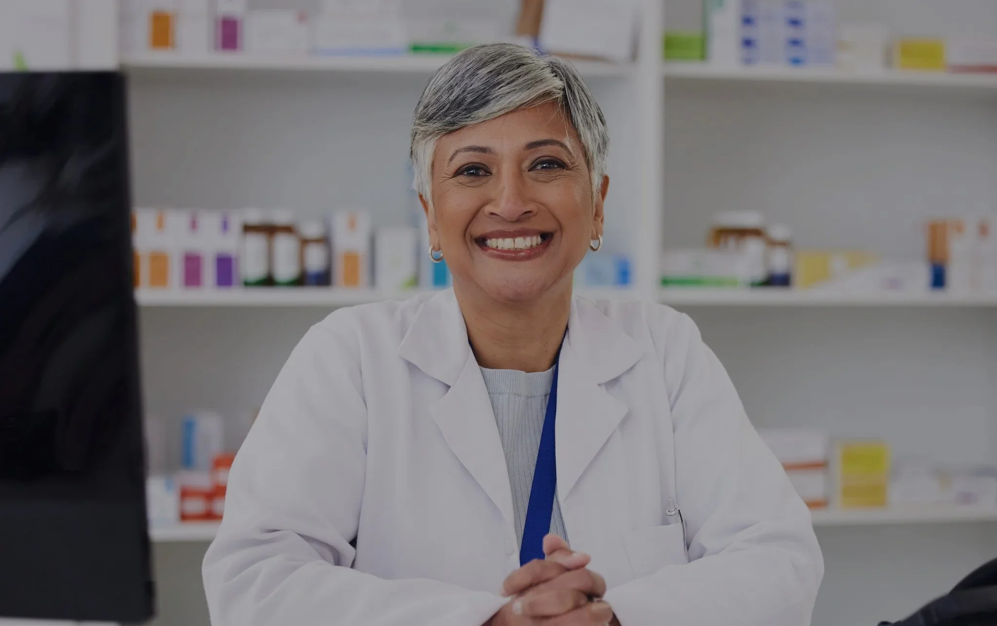 A smiling female healthcare professional with short gray hair, wearing a white lab coat, sitting at a desk in a medical office or pharmacy with shelves of medication bottles and boxes in the background.