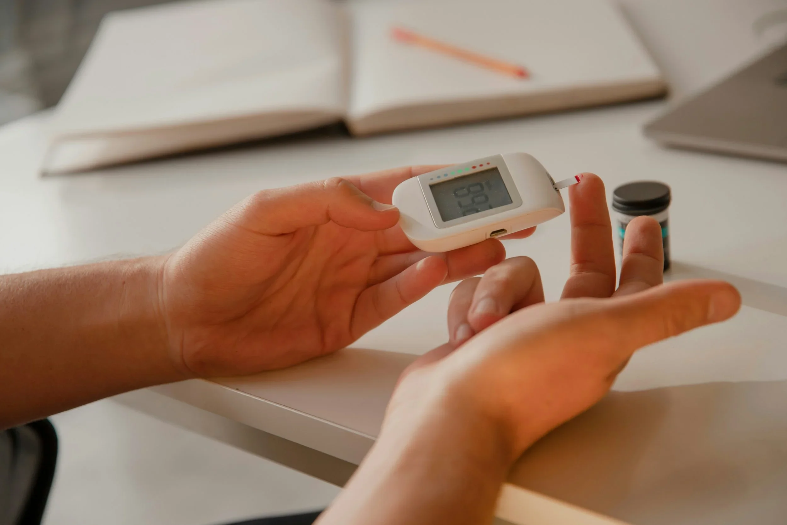Person testing blood sugar level using a digital glucose meter and a test strip, with a container of test strips on a white table.