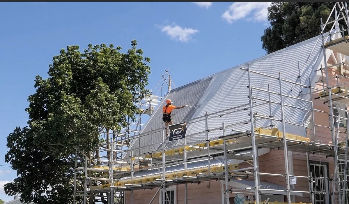 A worker wearing an orange safety helmet and vest cleaning or inspecting the metal roof of a house from scaffolding.