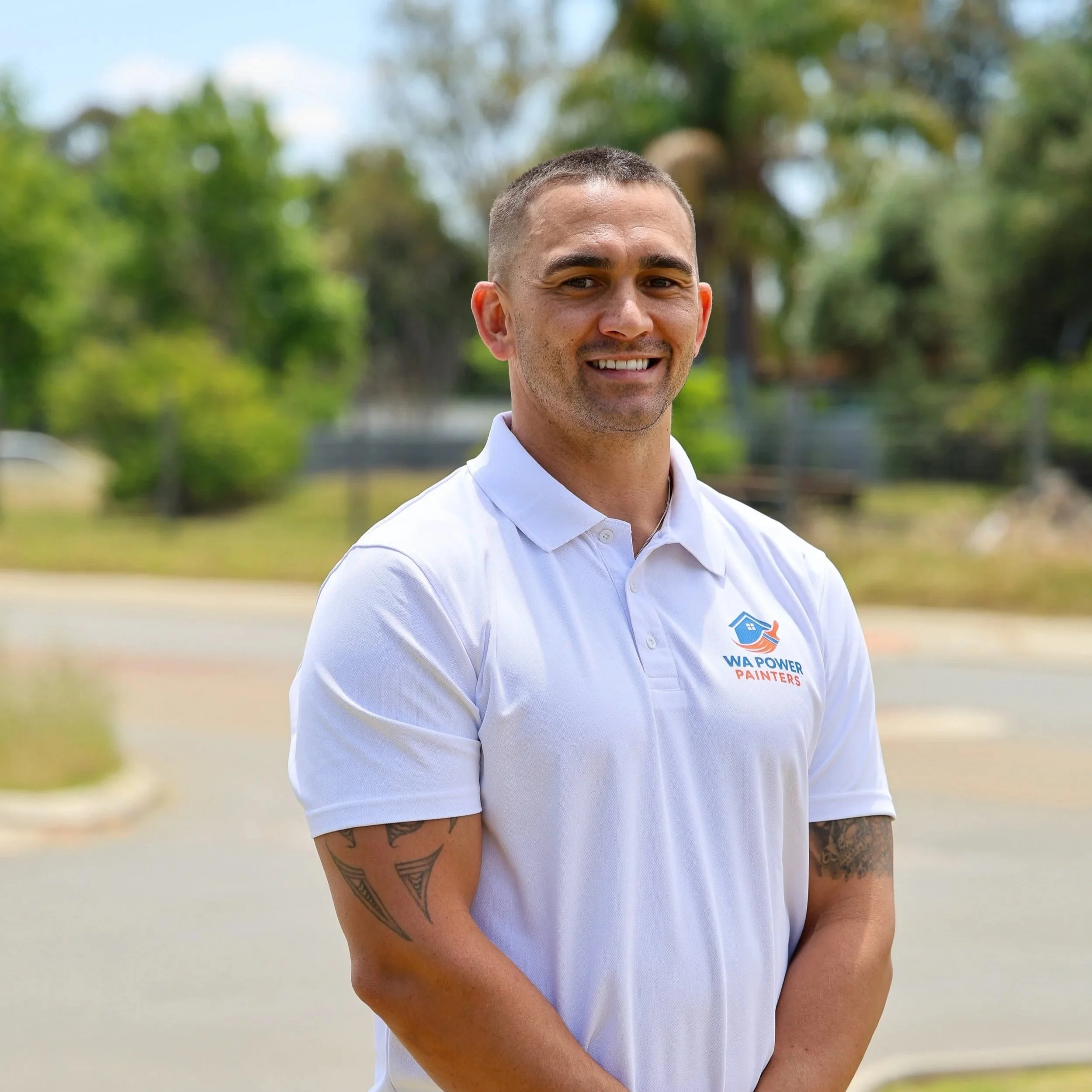 A smiling man in a white polo shirt with a 'WA Power Painters' logo, standing outdoors with trees and a cloudy sky in the background.