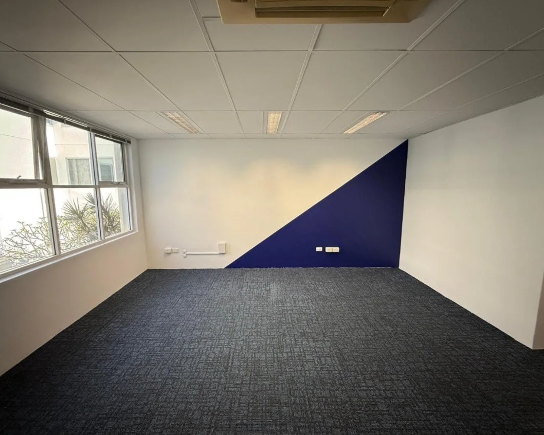 Empty office room with a window, gray carpet, white walls, and a blue geometric accent wall.