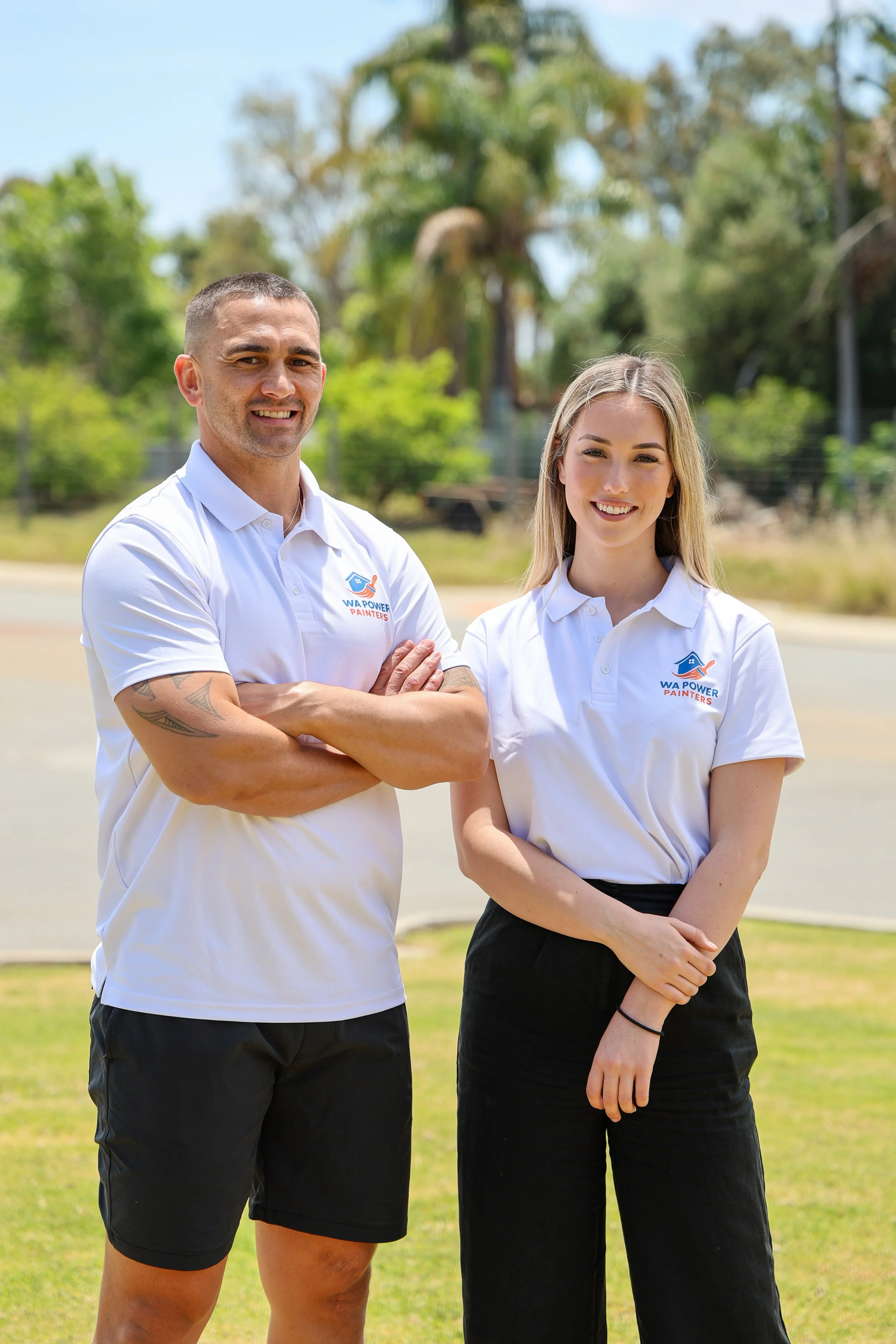 Two people standing outdoors on a sunny day, both wearing white polo shirts with a logo for WA Power Painters. The man has short hair, tattoos on his arms, and is smiling with arms crossed. The woman has long blonde hair, and is also smiling with one arm crossed over her waist.