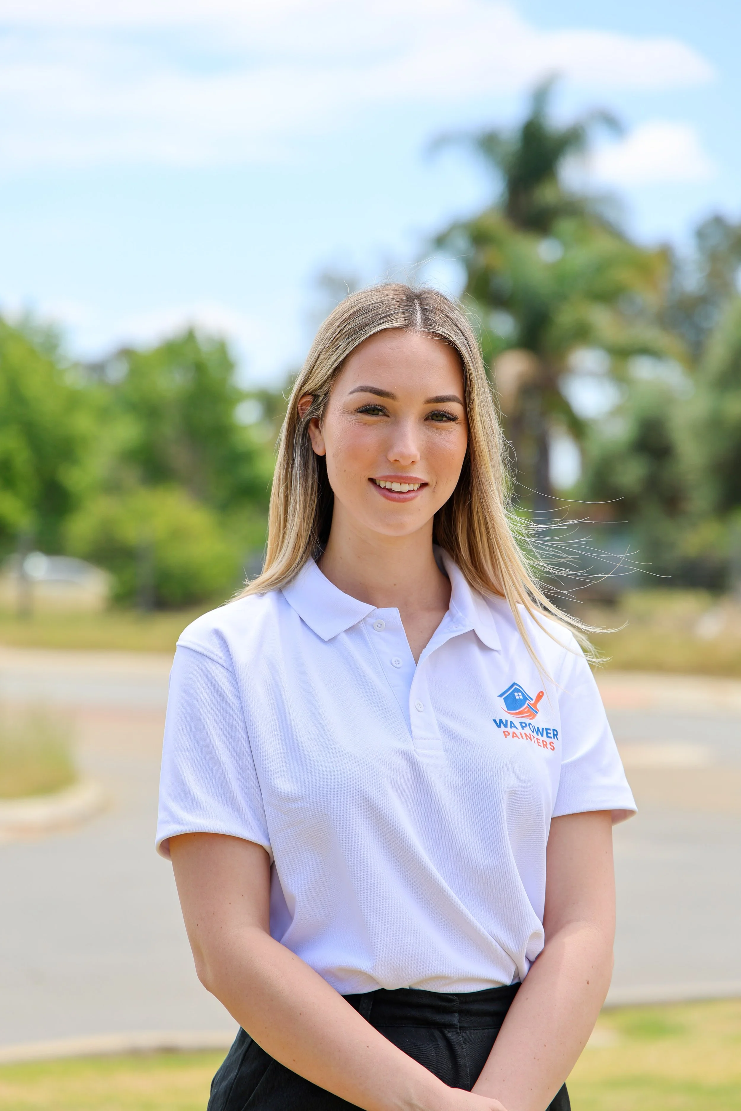 A young woman with long blonde hair, wearing a white polo shirt with a logo and black pants, standing outdoors with trees and a blue sky in the background.