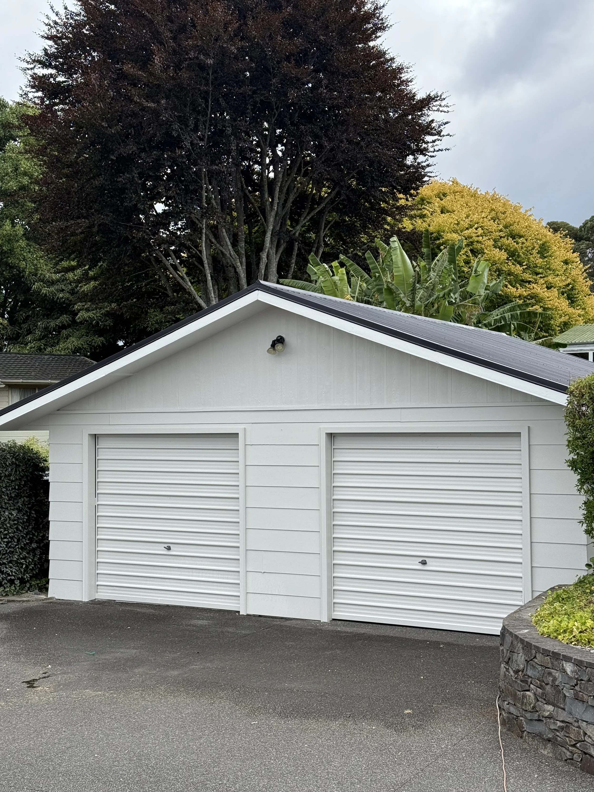 A white garage with two closed roll-up doors, a sloped roof, and a small light fixture above the doors. There are large trees and bushes in the background and around the garage.