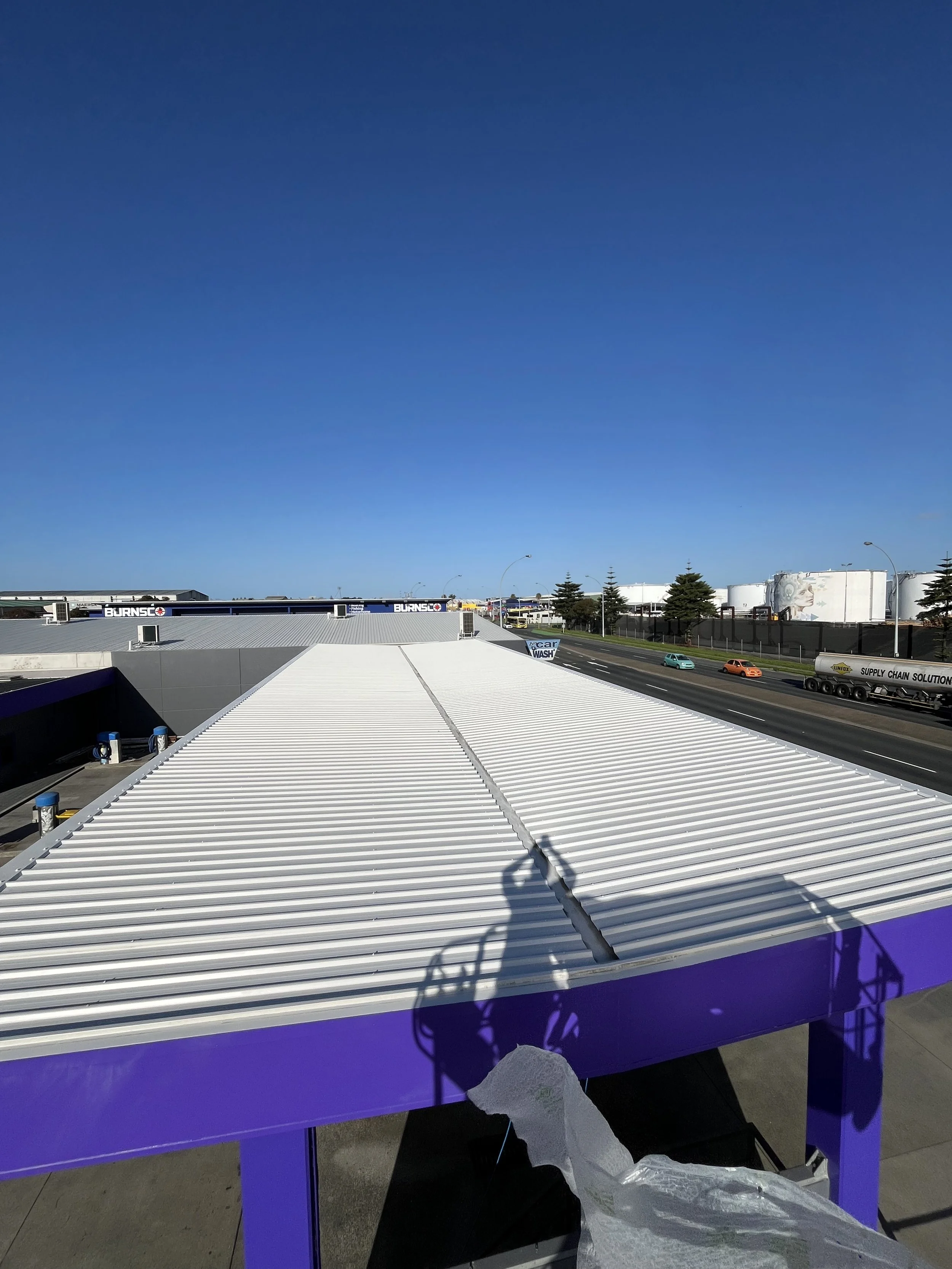 View of a roof with white metal panels and purple supporting structure, shadows cast by a crane, on a clear day with blue sky; in the background, a street with moving cars and industrial buildings.