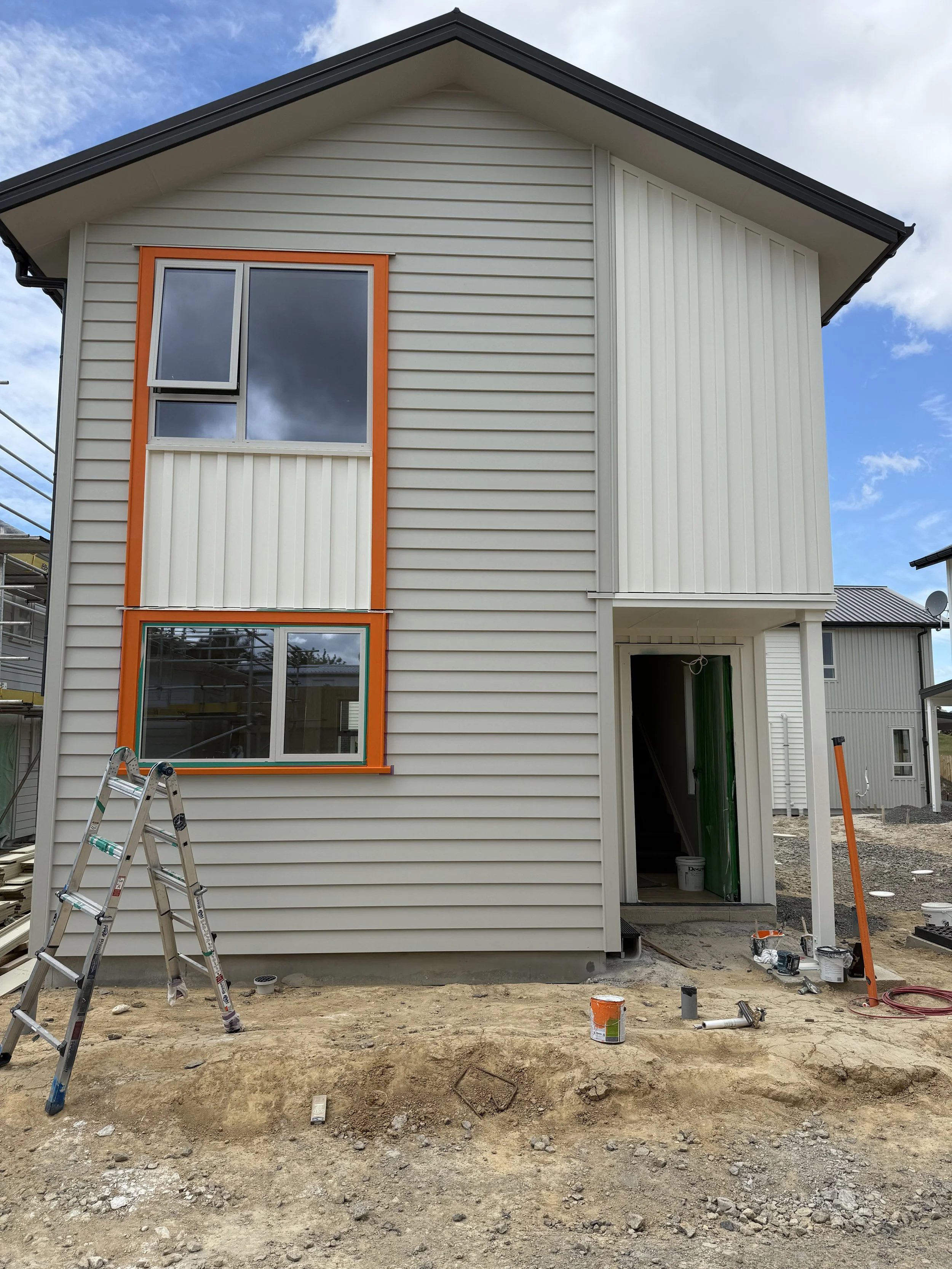 Under construction two-story house with white siding, orange window framing, and a front entryway under construction, with tools and materials on the ground.