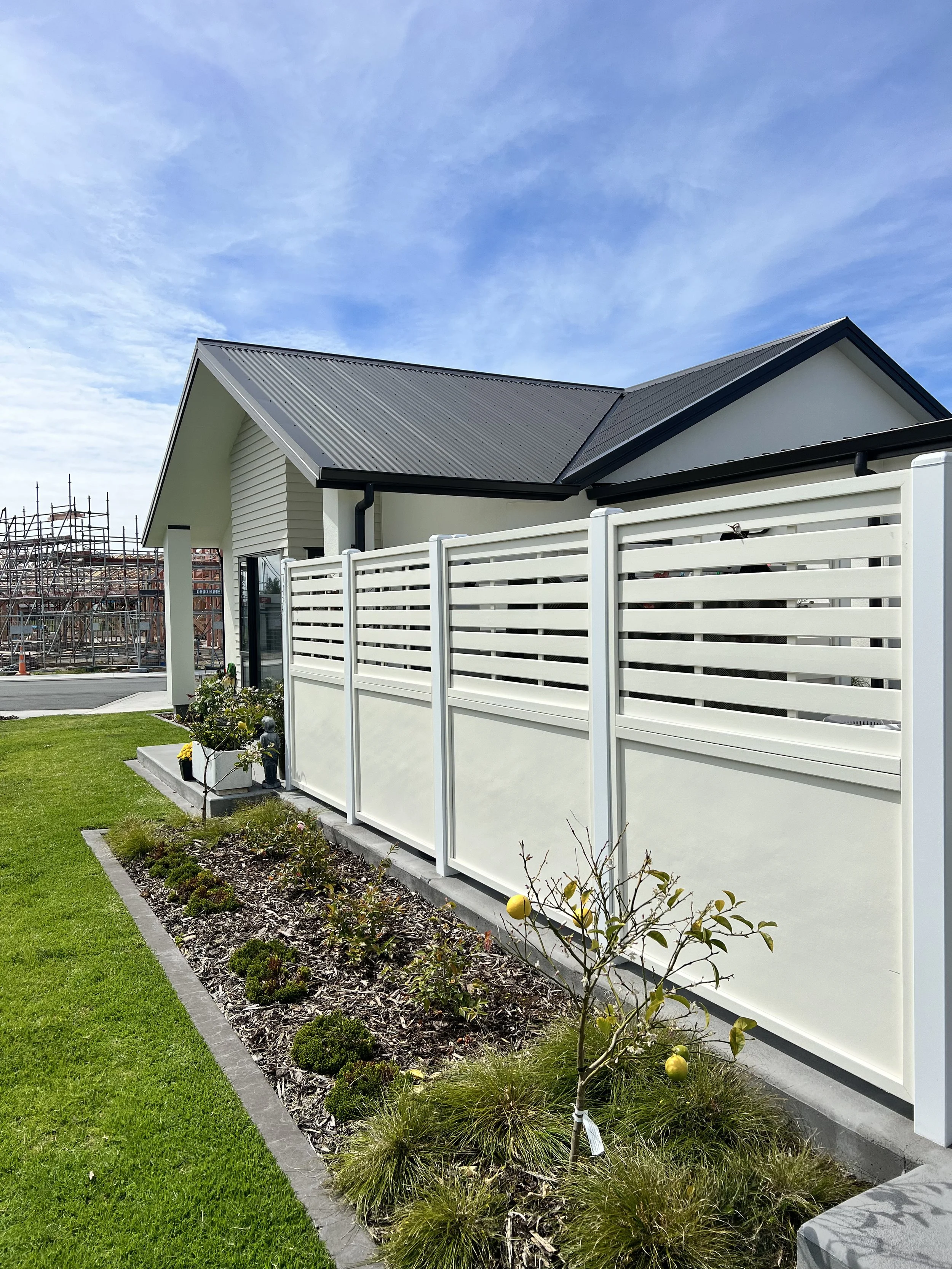 View of a modern house with a white privacy fence, a small garden with plants and a young fruit tree, and a gravel driveway under a partly cloudy sky.