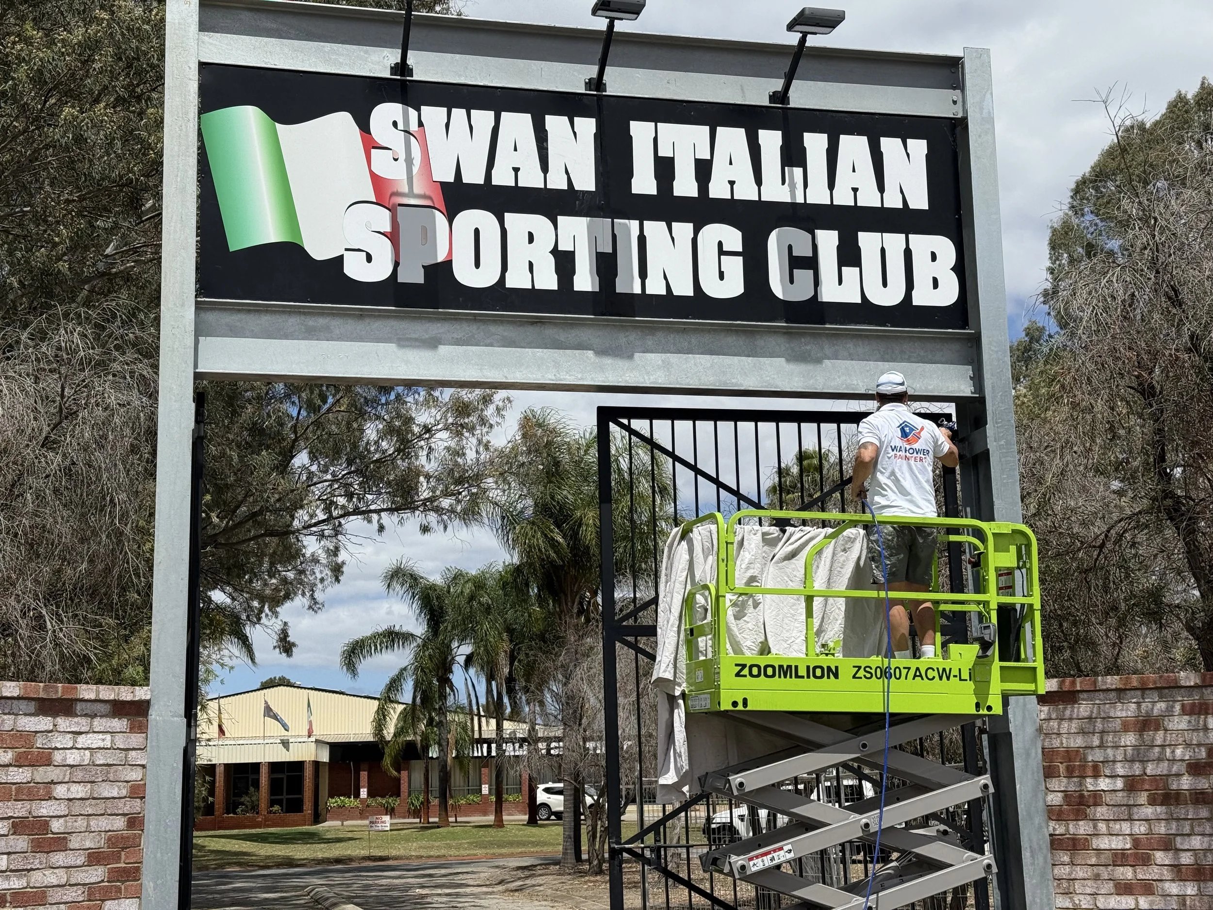Worker on a yellow scissor lift installing or maintaining a large sign for the Swan Italian Sporting Club, with trees and a building in the background.
