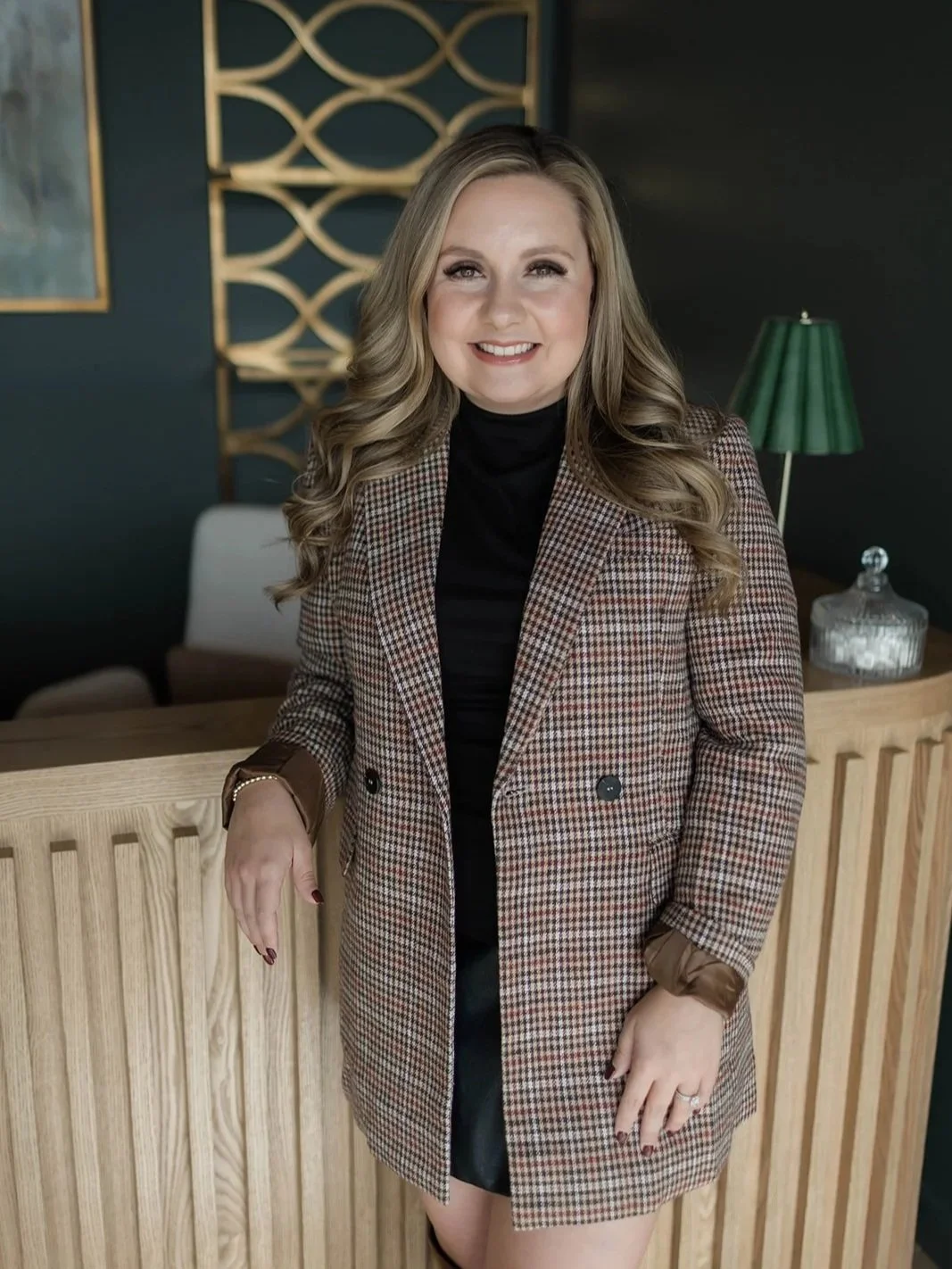 A woman with long, wavy blonde hair smiling, standing indoors at a wooden bar or counter, wearing a houndstooth blazer over a black top, with a green lamp and decorative wall art in the background.