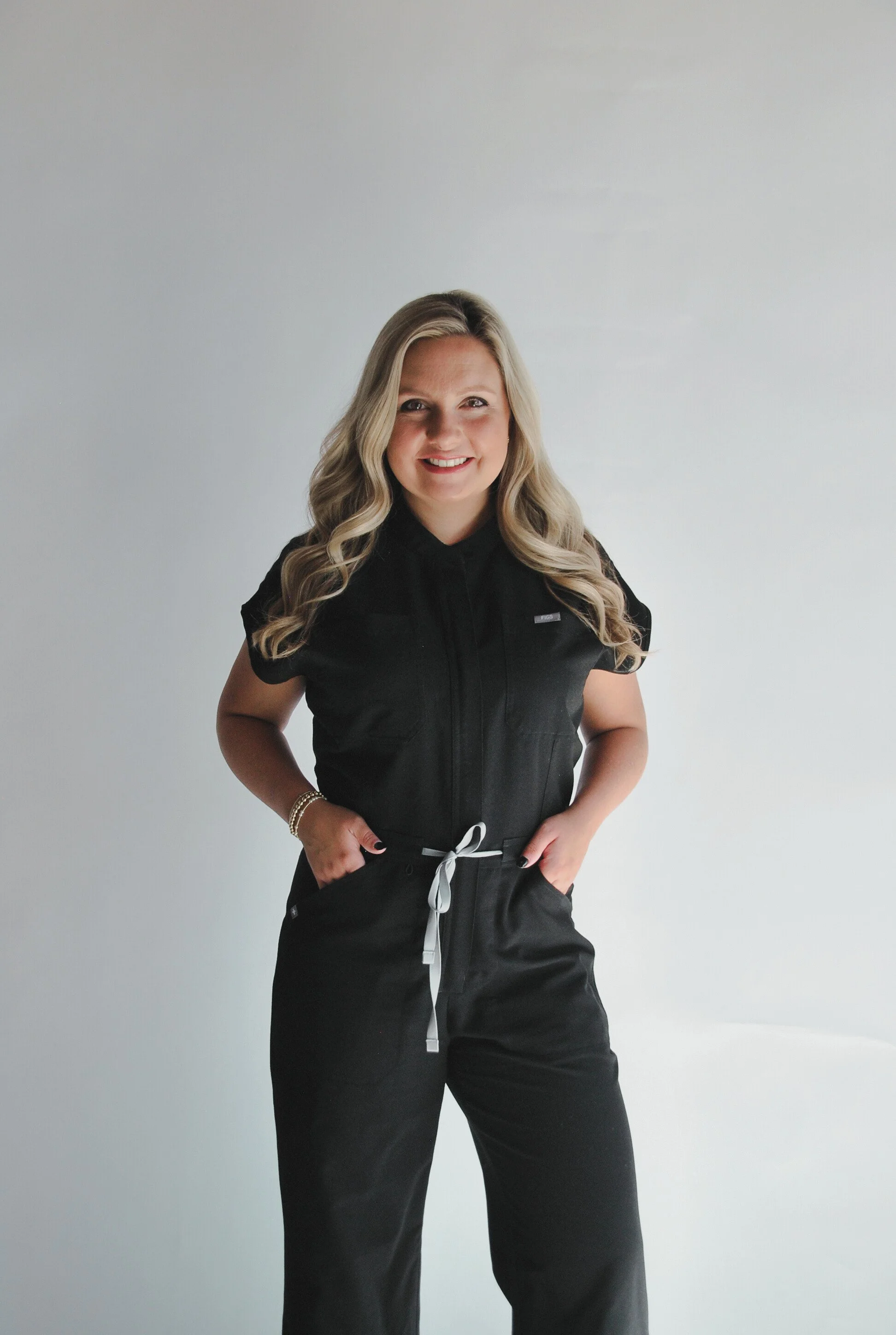 A woman with long blonde hair smiling and wearing a black jumpsuit standing against a plain white wall.
