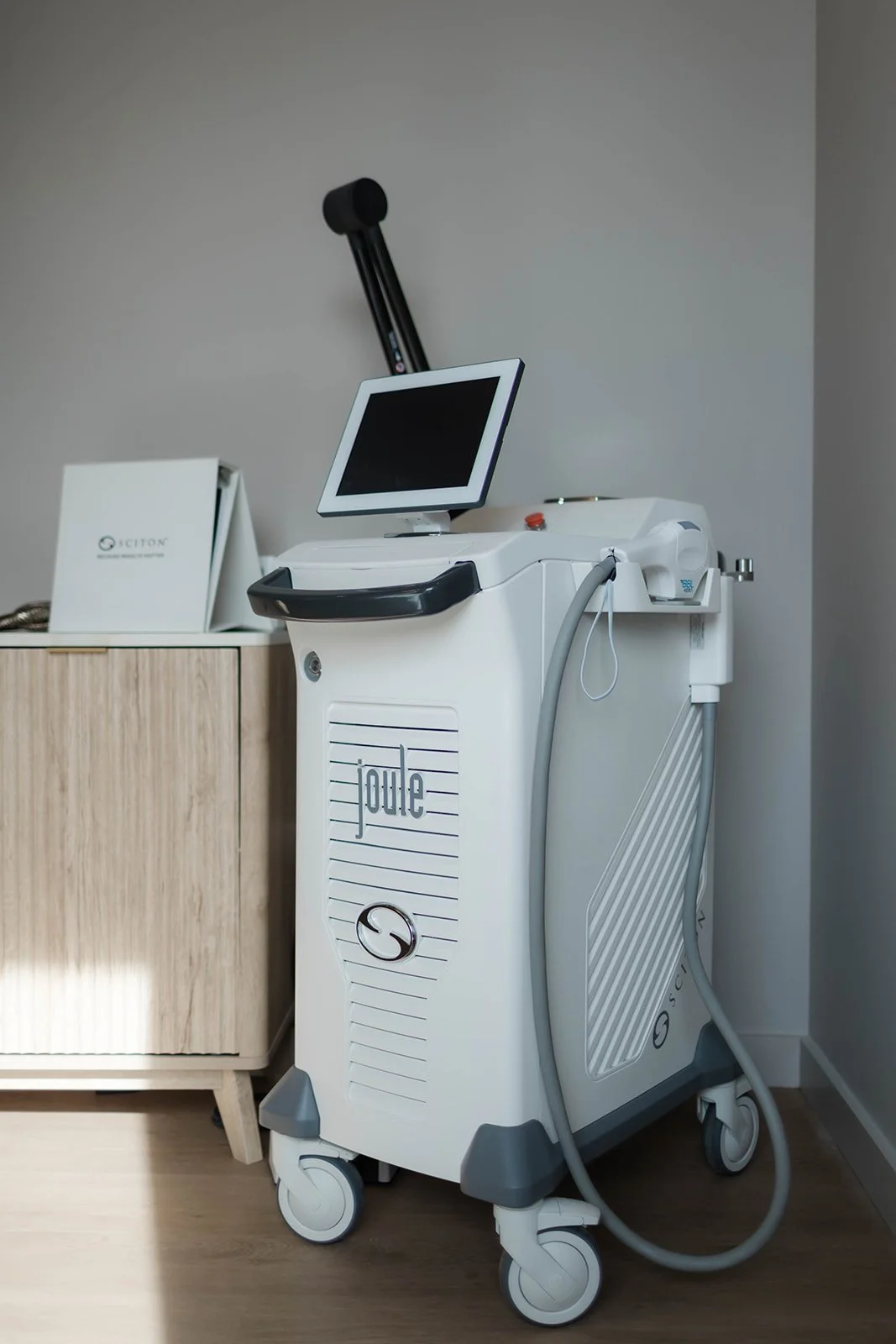 Medical laser device with a touchscreen monitor, on wheels, placed beside a light wood cabinet in a clinical setting.