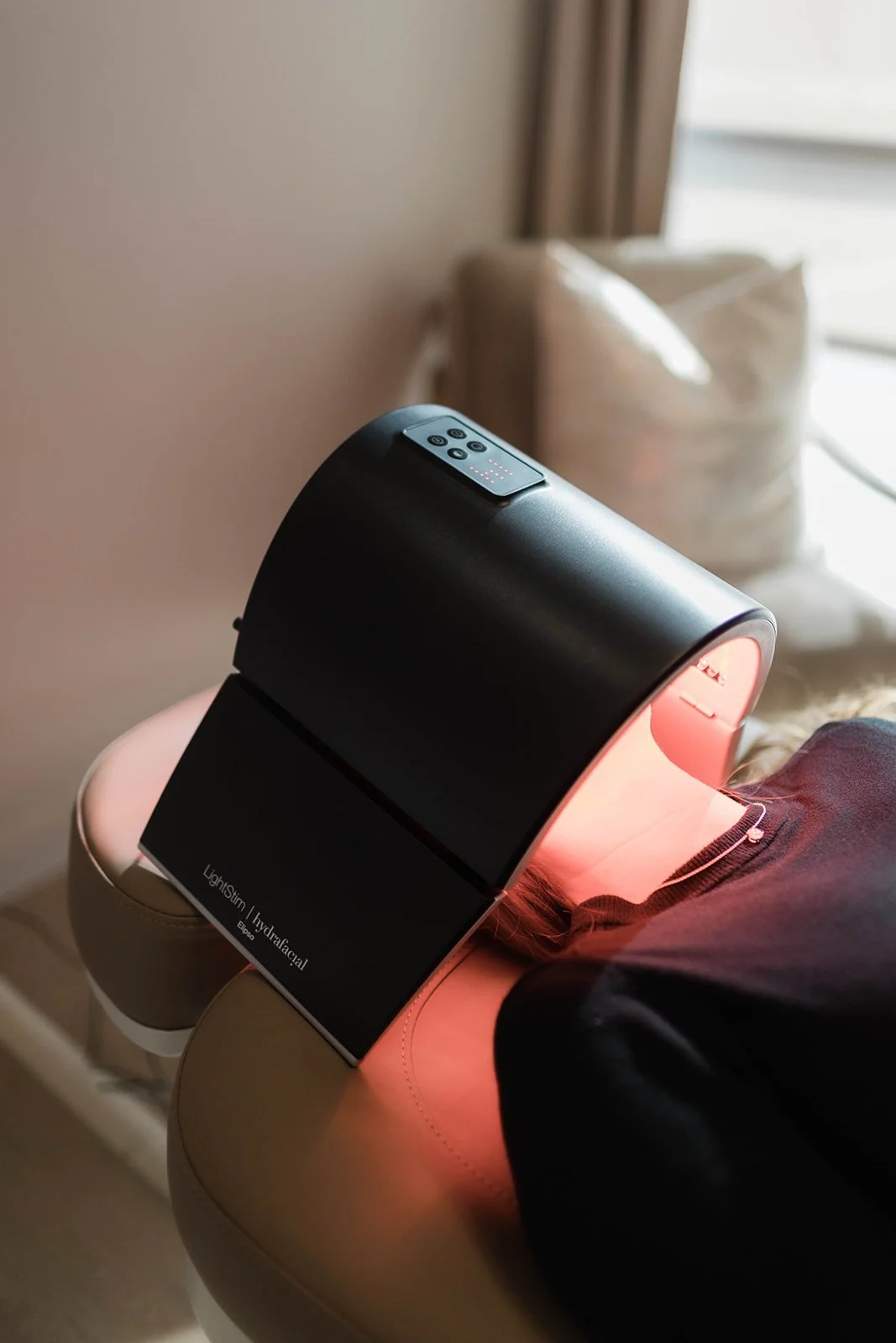 A person receiving a facial treatment with a red light therapy device on their face, in a spa or skincare clinic setting.