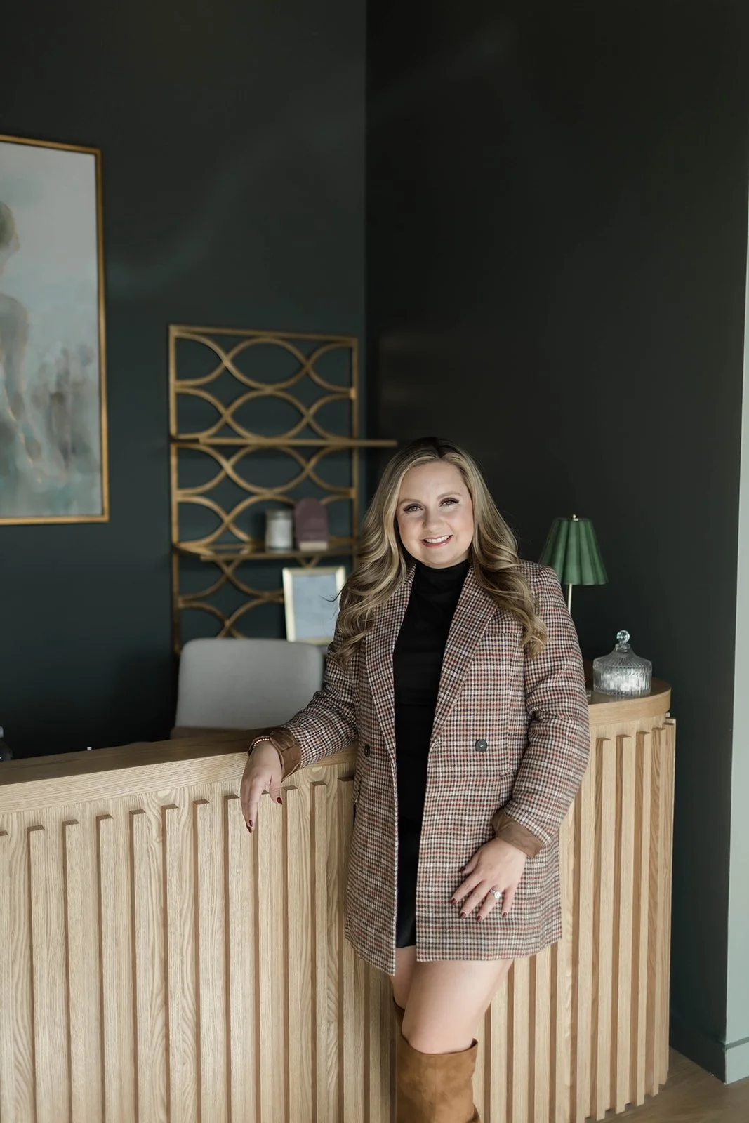 A woman with long blonde hair smiling, standing next to a wooden reception desk. She is wearing a brown plaid blazer, black turtleneck, and brown knee-high boots, in a modern interior with dark walls, a green lamp, and decorative shelves.
