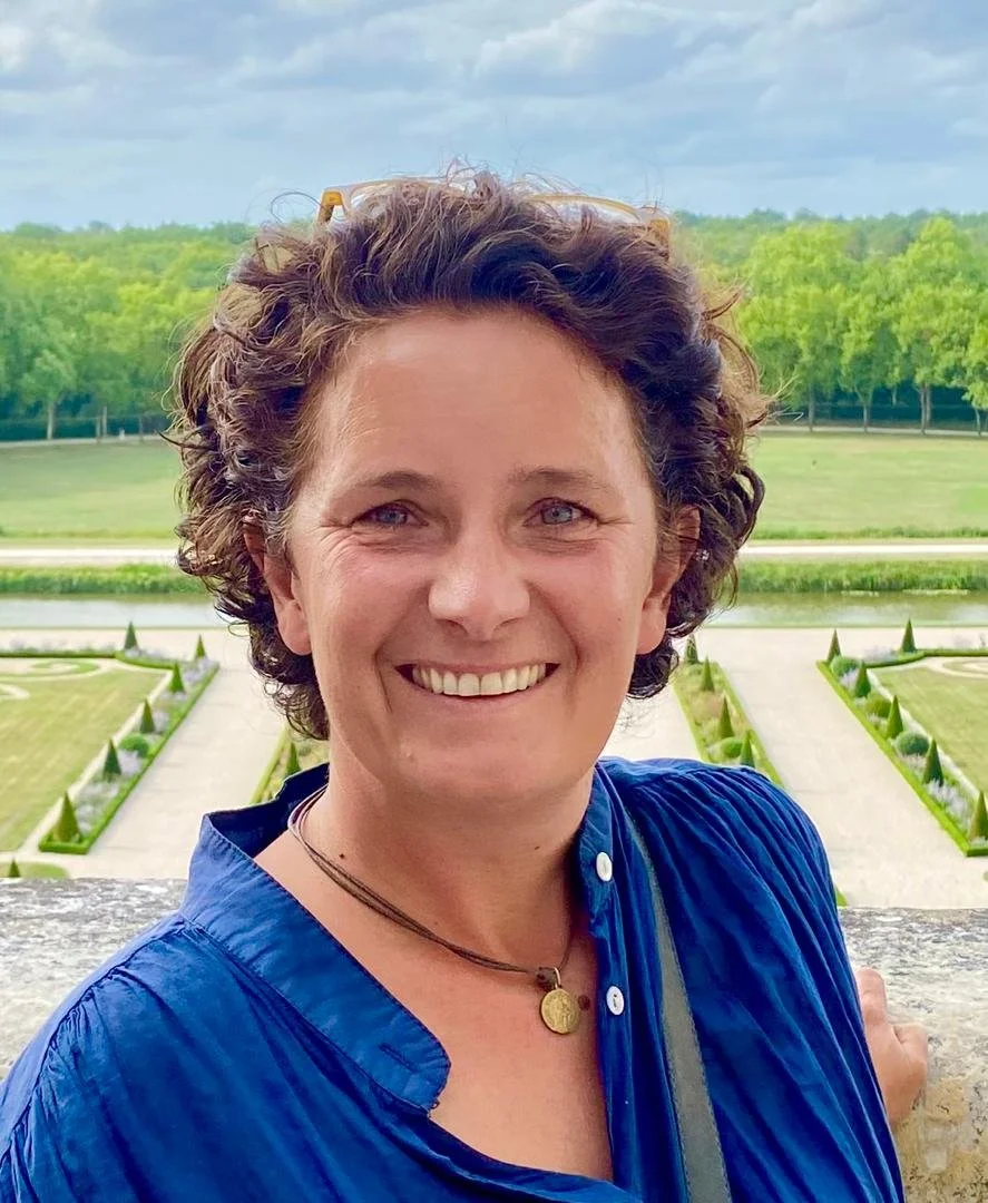 A smiling woman with curly brown hair, wearing a blue shirt and glasses on her head, standing outdoors with a garden and trees in the background.