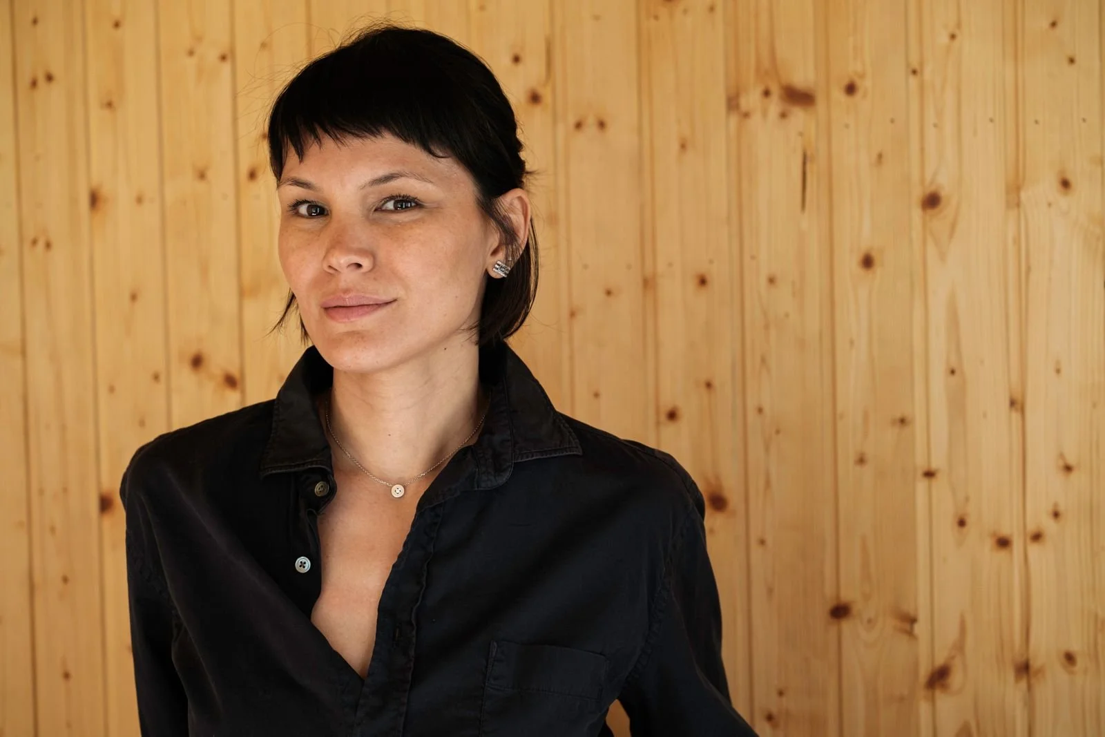 A woman with short black hair wearing a black shirt and jewelry, standing against a wooden panel background.