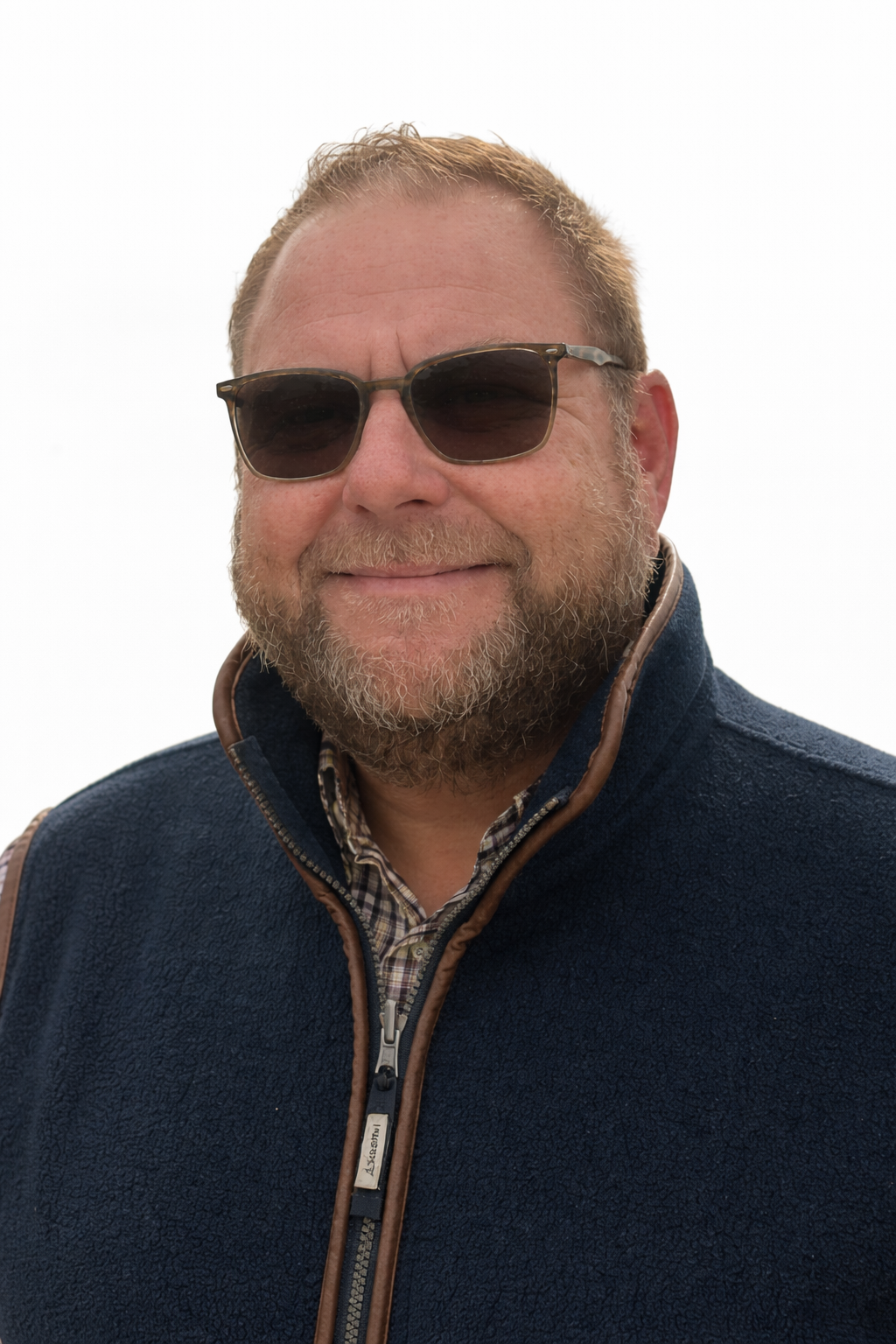 A man with a beard wearing sunglasses, a navy blue jacket with brown trim, and a plaid shirt underneath, smiling against a white background.