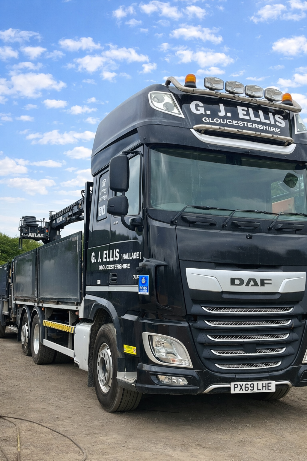 A black DAF truck with the company name G. J. Ellis Gloucestershire written on the front and side, equipped with a crane, parked on a gravel surface under a partly cloudy sky.