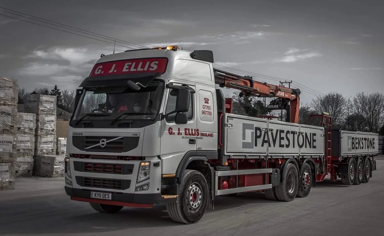 A large white flatbed truck with a crane attachment parked on a dirt surface, carrying pallets with stored items in the background.