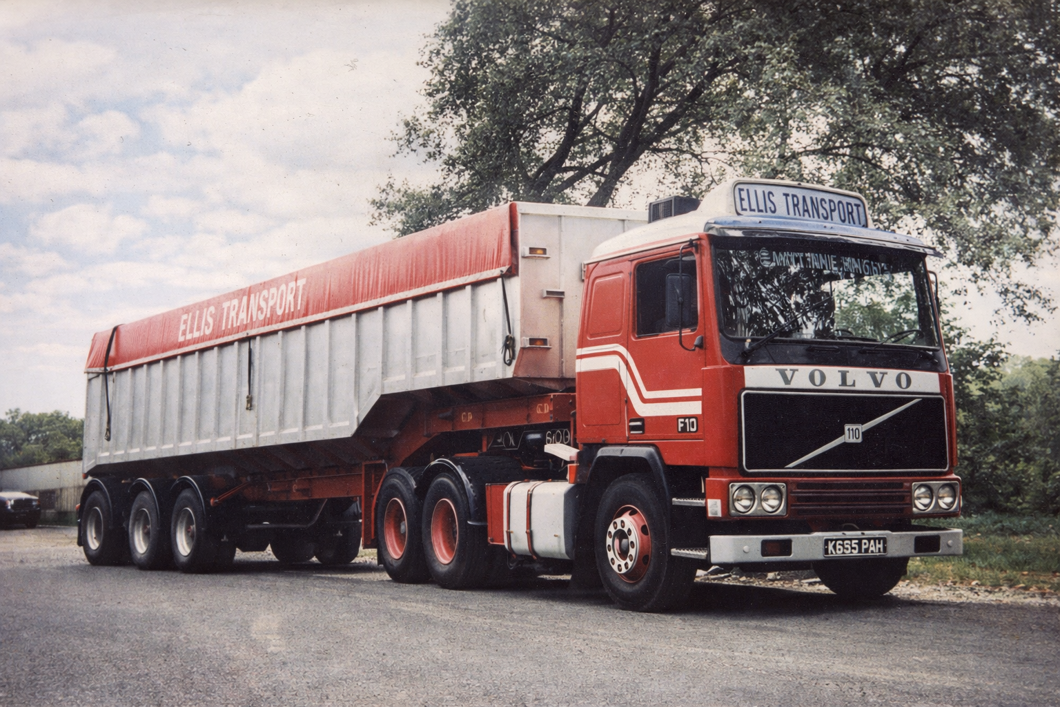 A vintage red Volvo transport truck with a white cargo container marked 'Ellis Transport,' parked on a gravel surface with trees and a partly cloudy sky in the background.