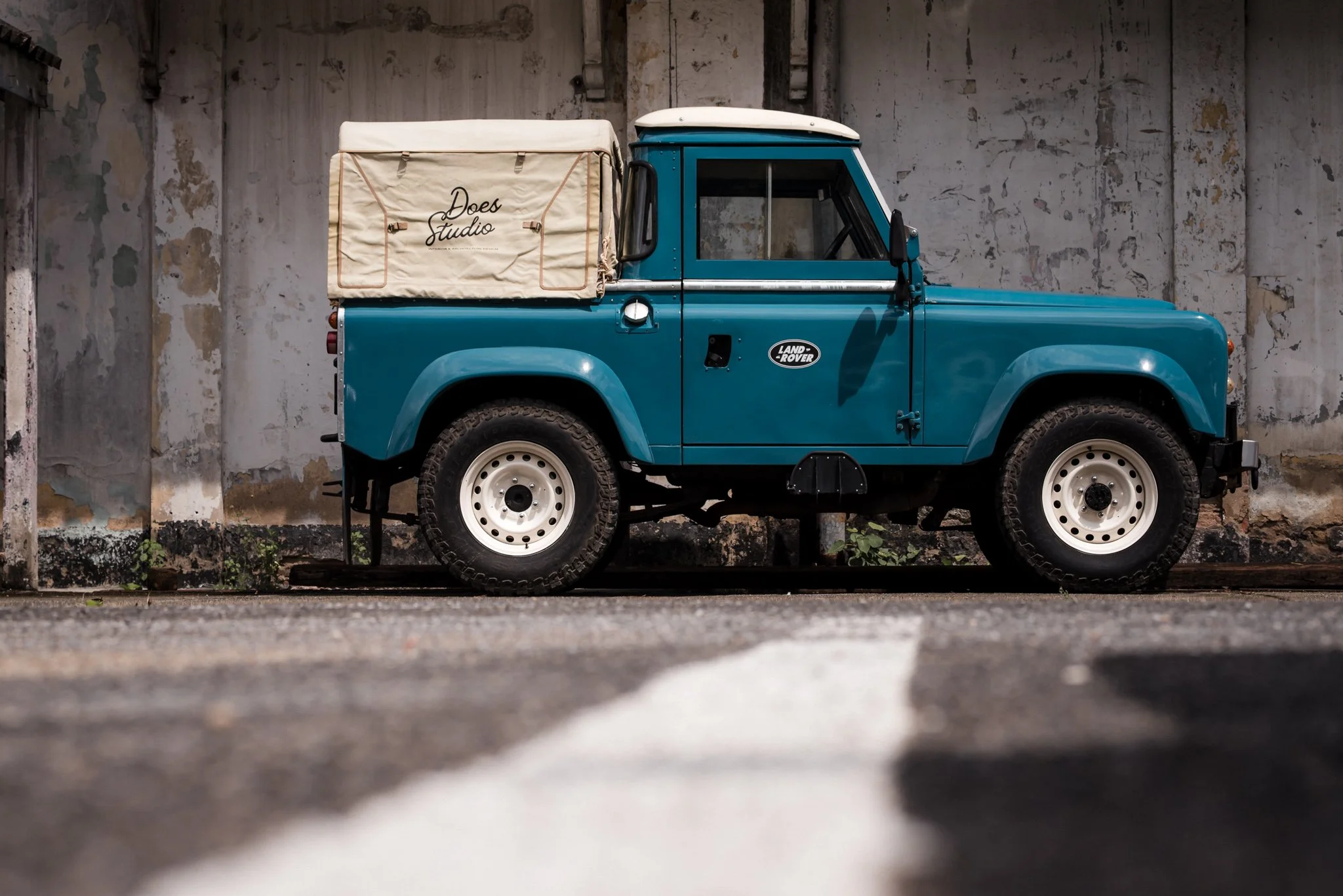 A vintage blue Land Rover parked against a weathered concrete wall with peeling paint.