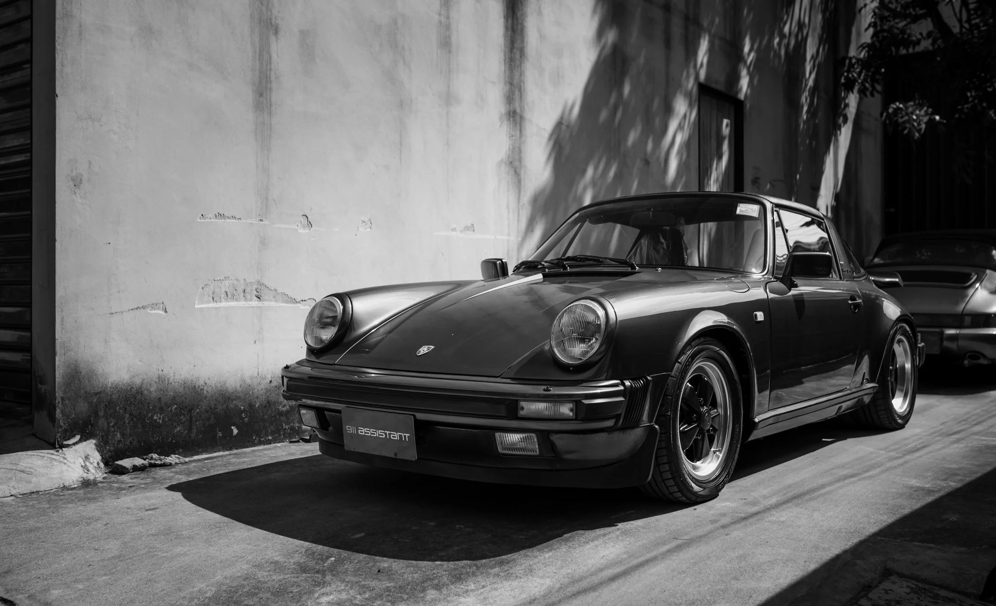 Black-and-white photo of a vintage Porsche 911 car parked beside a textured wall and a tree casting shadows.