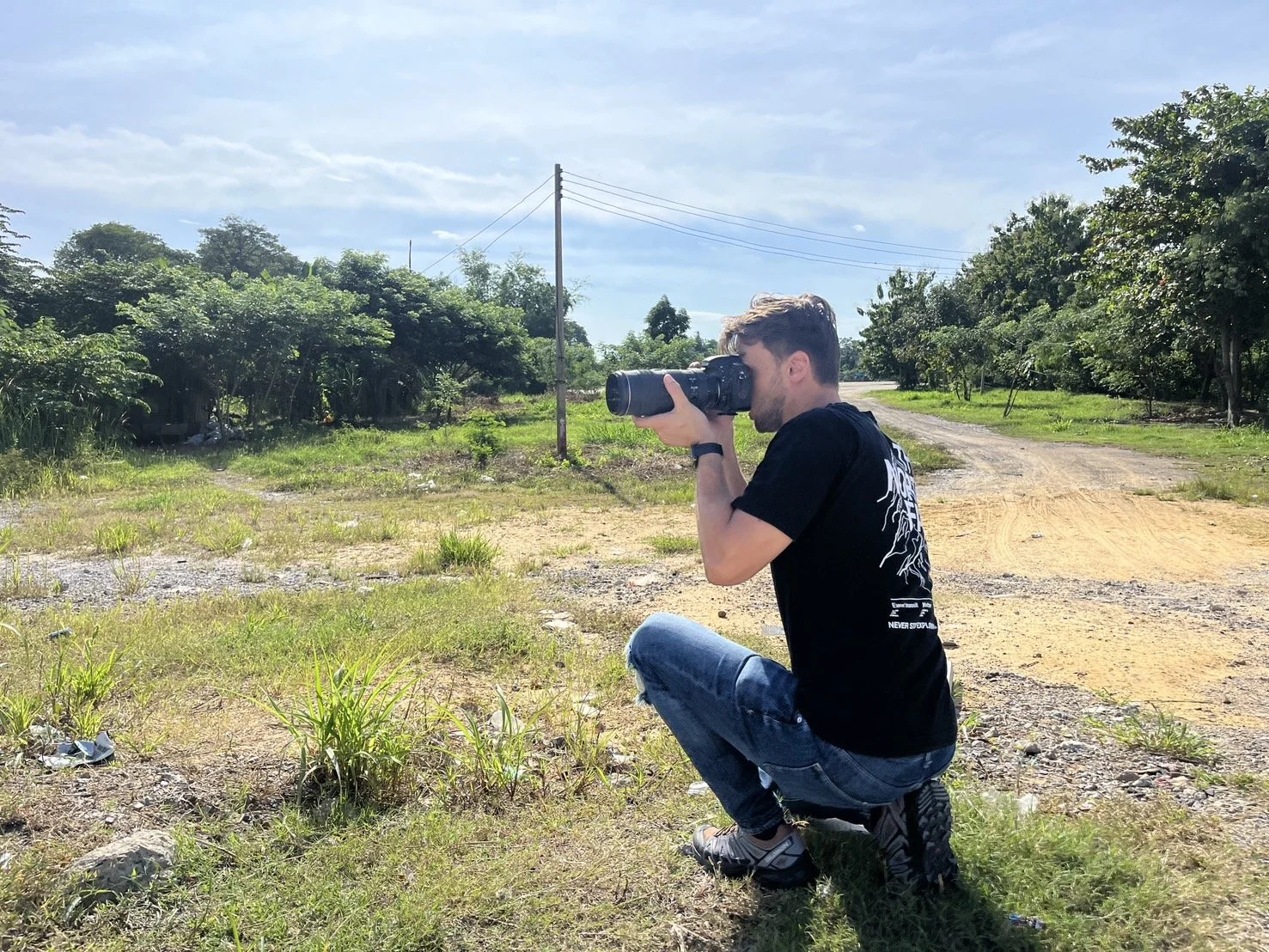 Arran Carter Cheetham, AJP Automotive squatting outdoors, taking photographs with a camera, on a sunny day with green trees and a dirt road in the background.