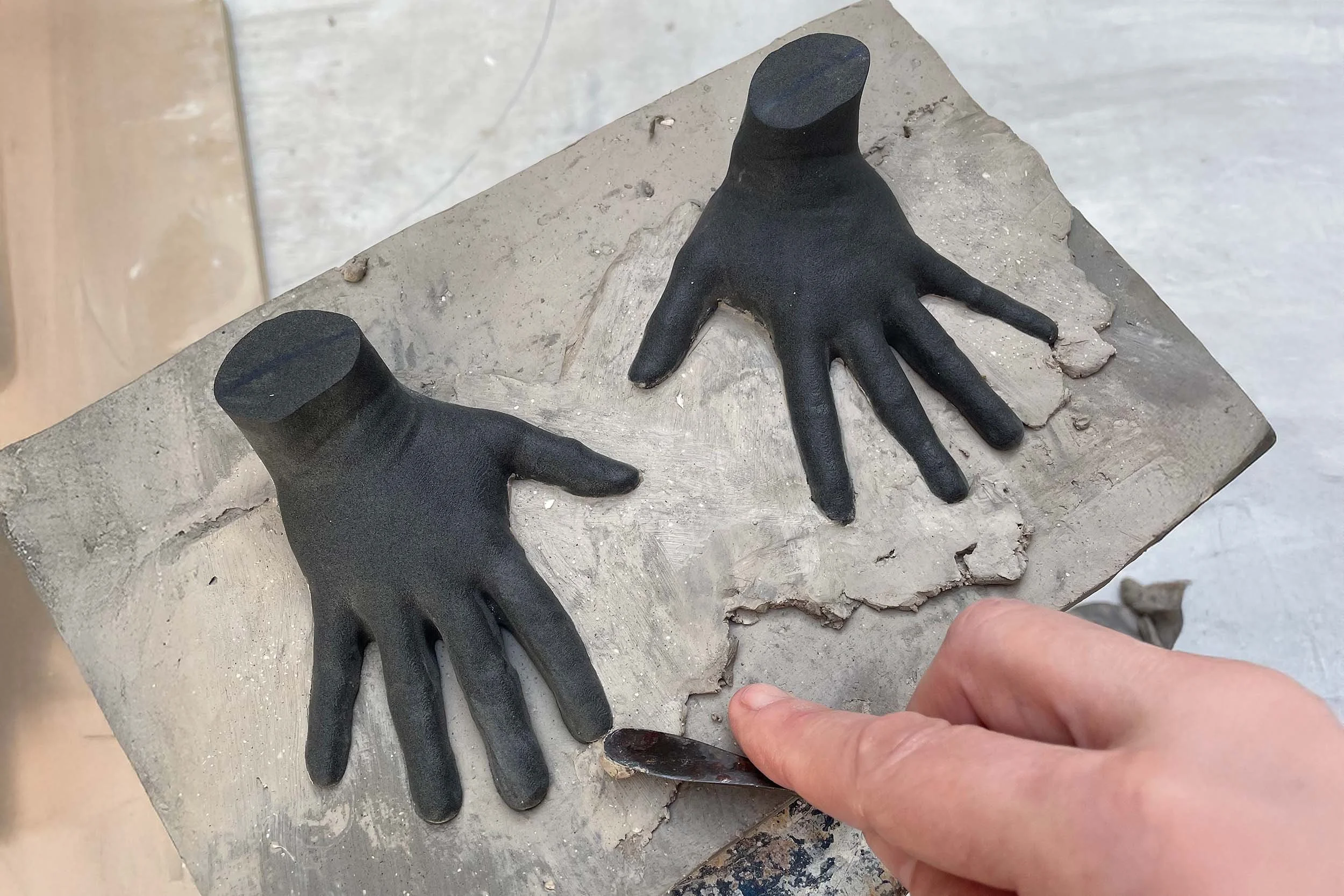 Two black plaster hands are mounted on a clay slab, with a person's finger pointing at the base of one of the hands.