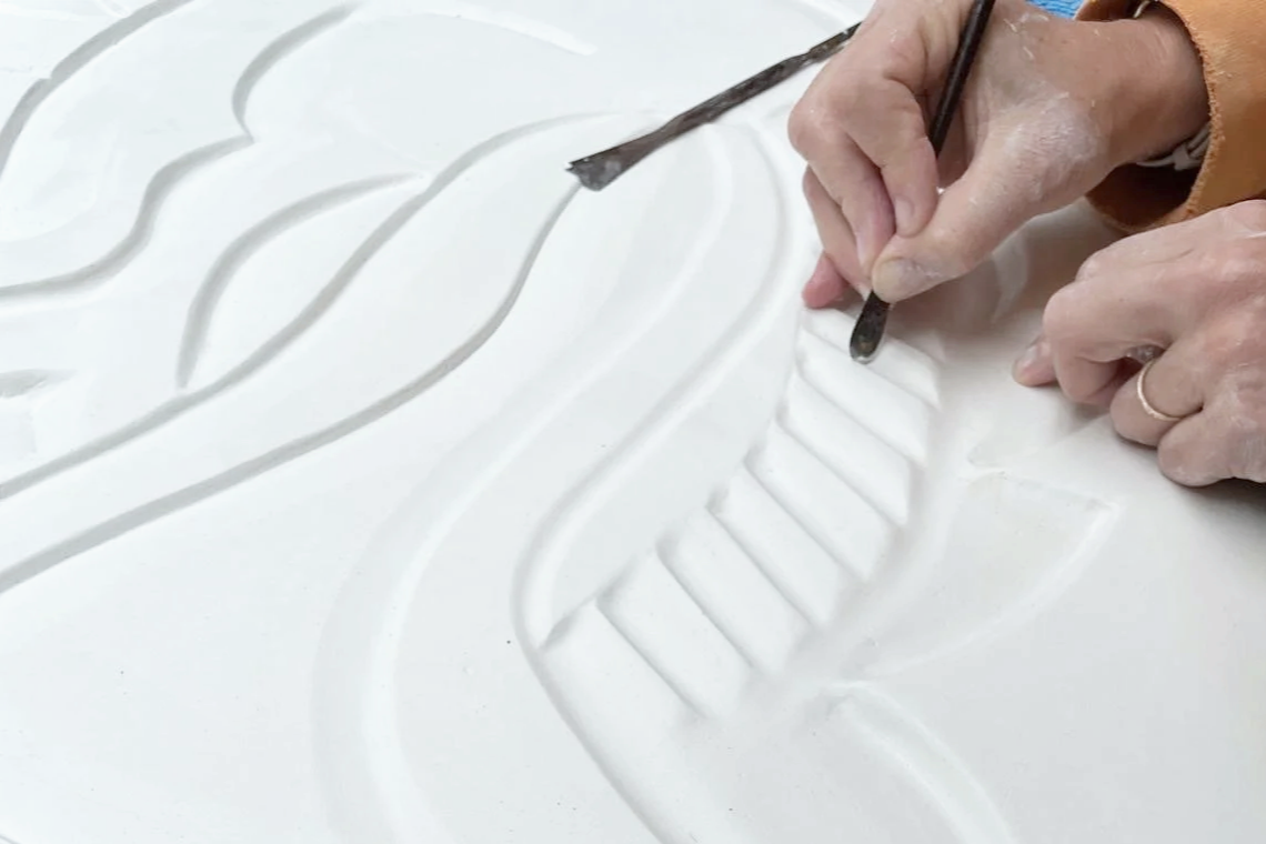 Person carving a detailed leaf design into a white plaster surface, with a small carving tool.