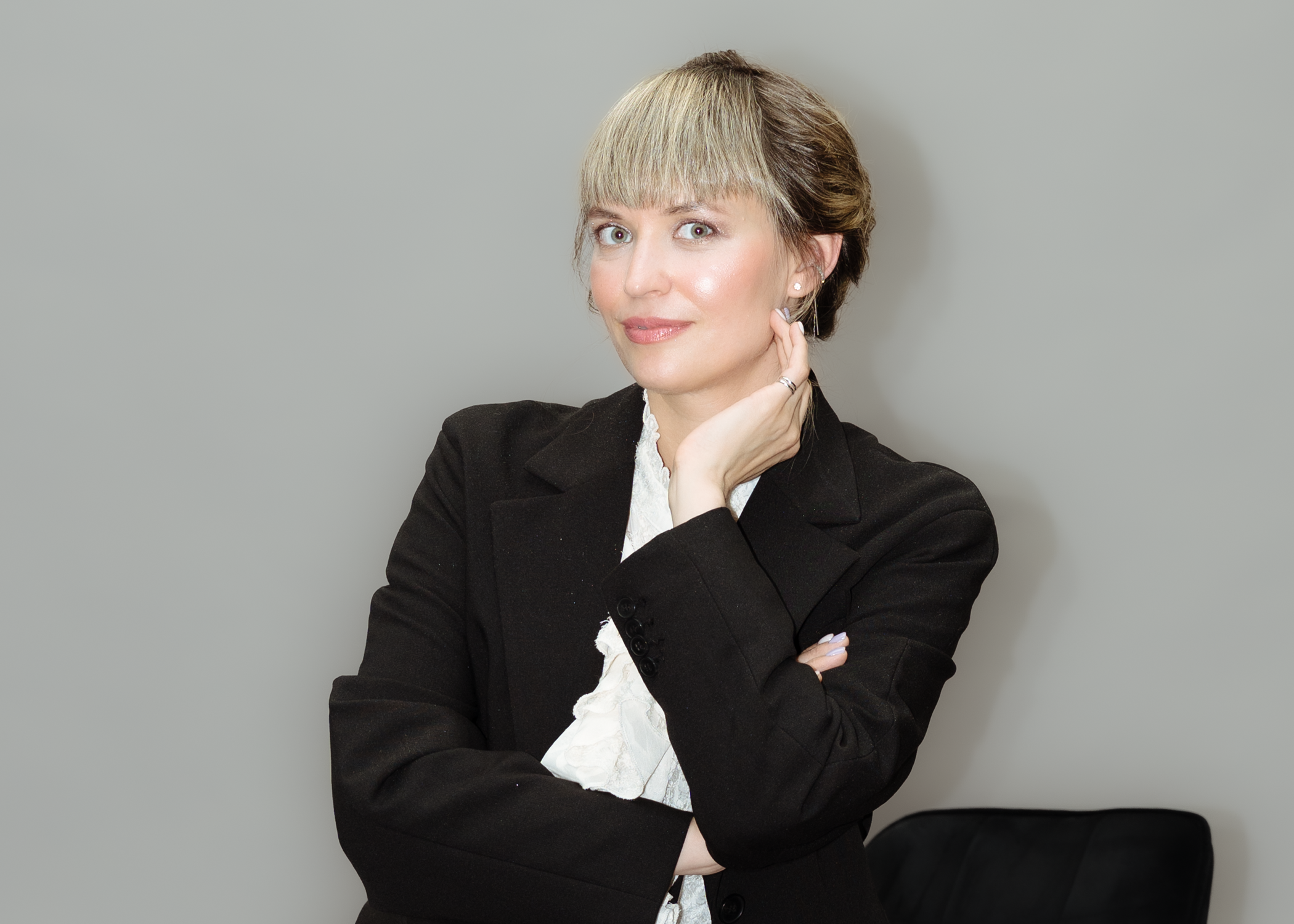 Eine Frau mit kurzen Haaren in einem schwarzen Blazer und weißer Bluse, die an einer Wand steht, lächelnd in die Kamera schaut, die Hand am Hals hält und eine Hand vor den Bauch verschränkt.