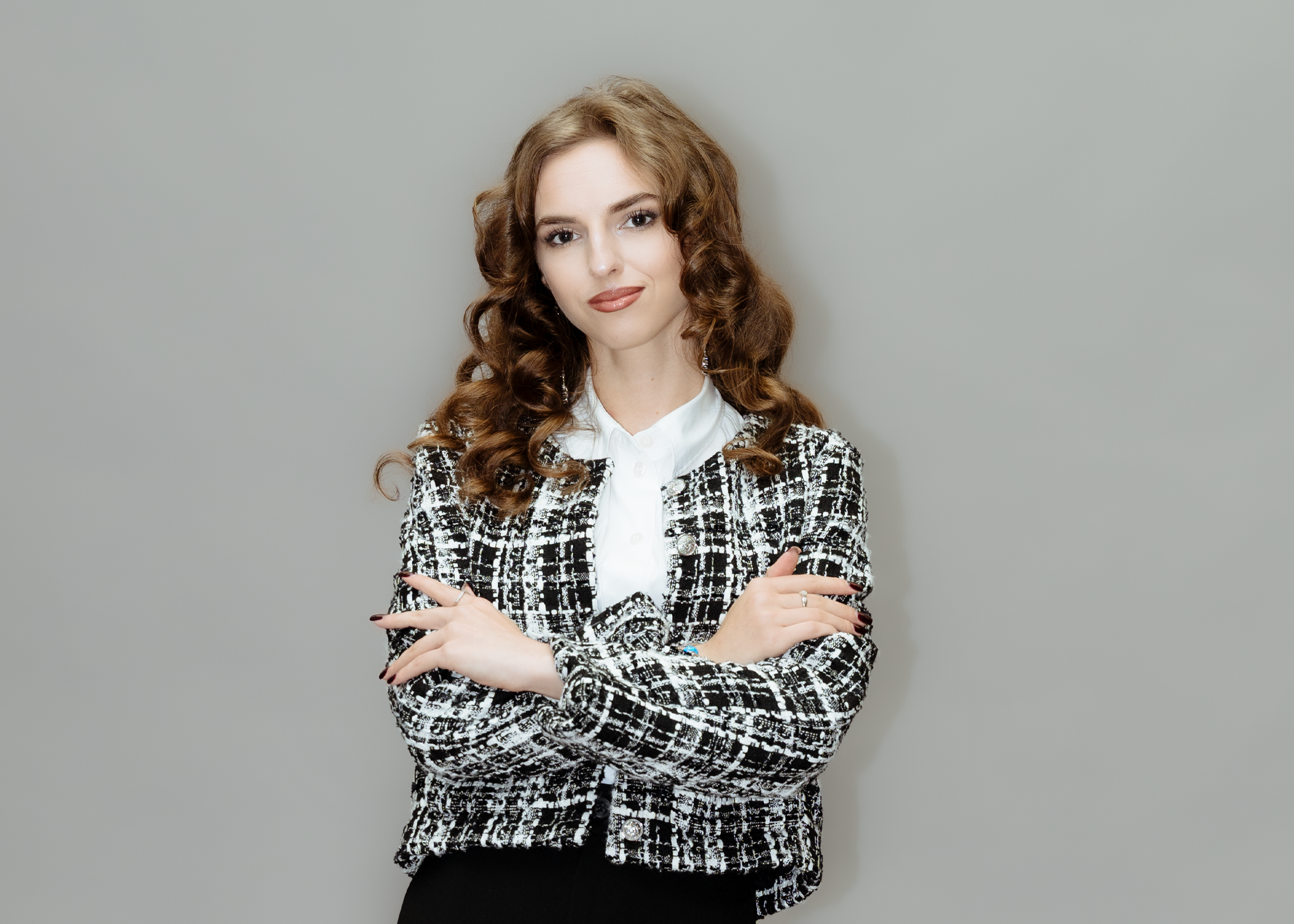 Eine Frau mit langen, lockigen, braunen Haaren, die vor einem grauen Hintergrund steht, trägt eine schwarz-weiße kariert Jacke und eine weiße Bluse und lächelt leicht.