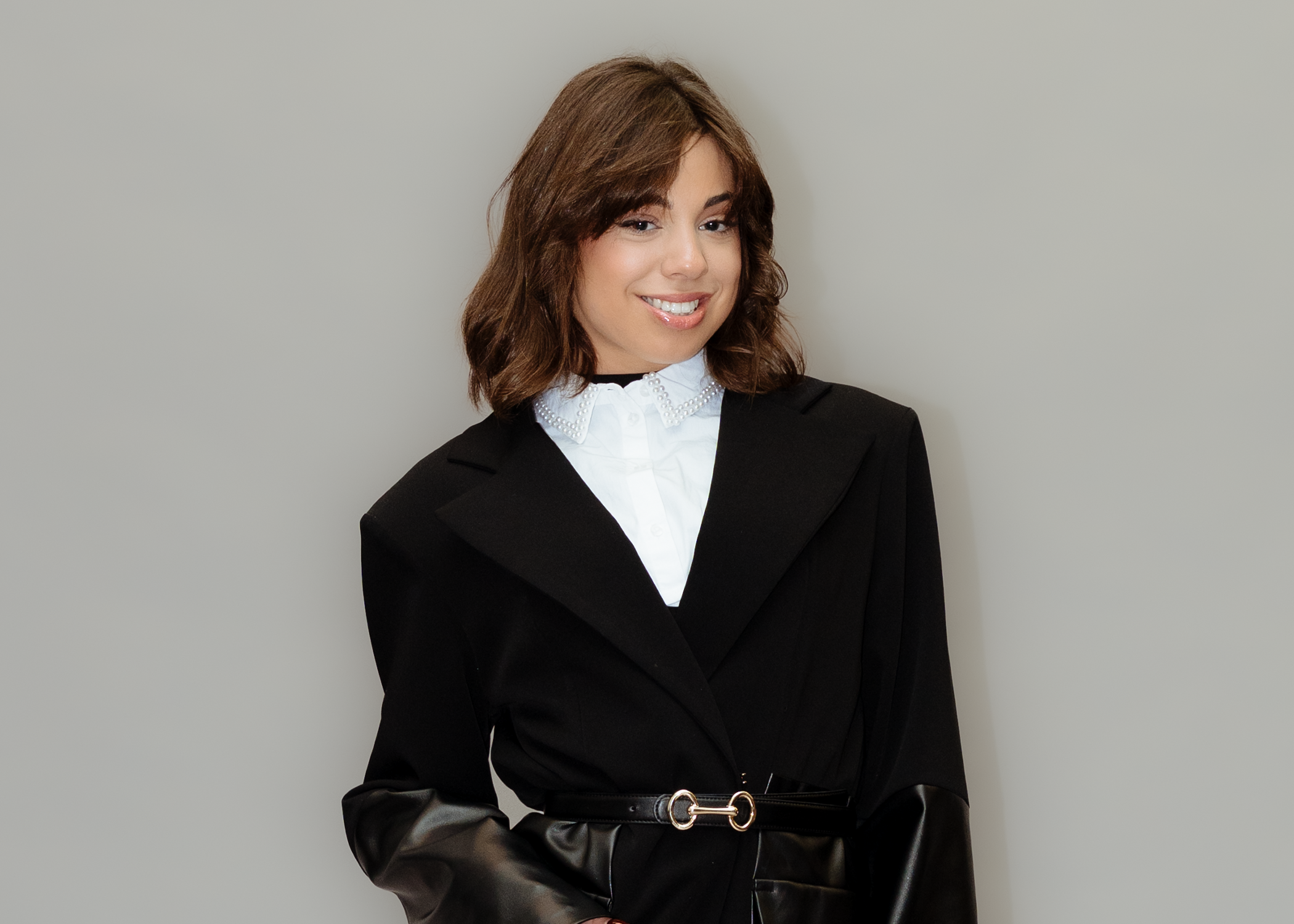 Frau trägt schwarze Jacke mit Lederärmeln und weißen Hemd mit Perlenkragen, lächelt vor hellem Hintergrund.
