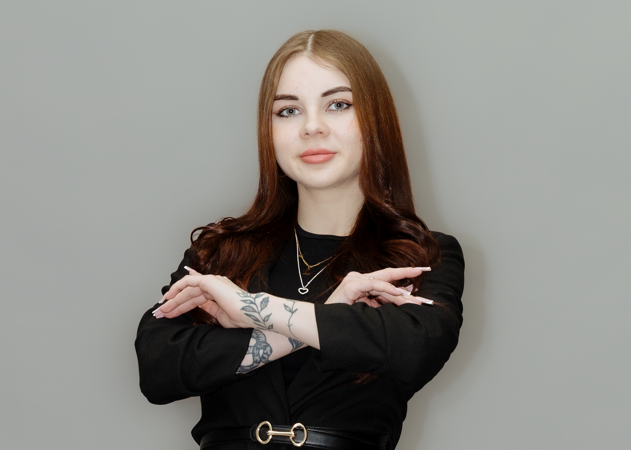 Eine junge Frau mit roten Haaren, sommern in einem schwarzen Blazer, trägt mehrere Halsketten und Tattoos an beiden Armen, steht vor einem hellgrauen Hintergrund.