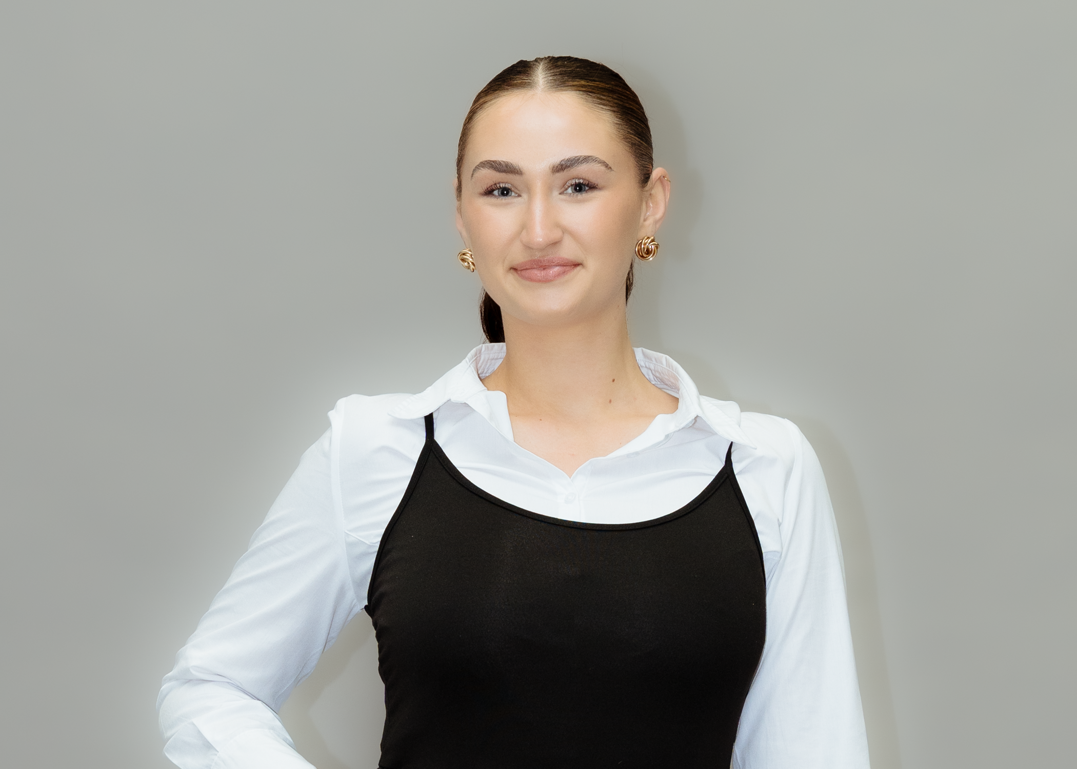 Eine junge Frau mit braunem Haar, die einen weißen Hemd und schwarzen Top trägt, posiert vor einem neutralen Hintergrund.