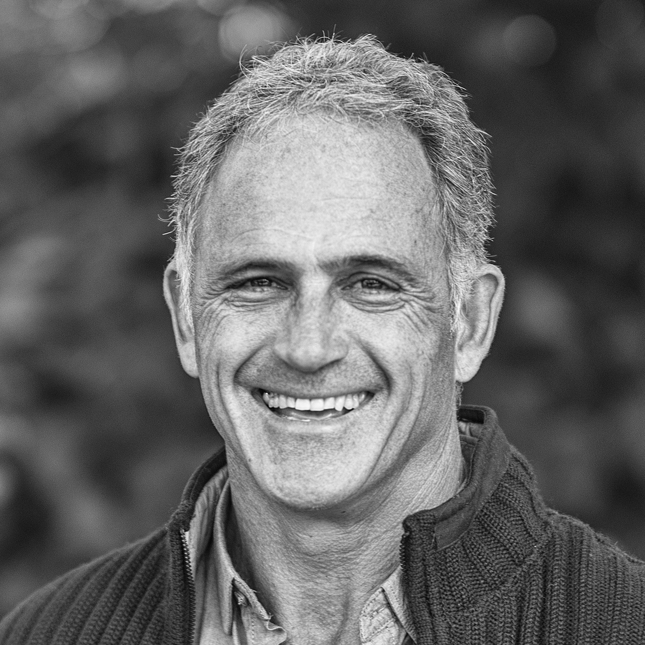 Black and white close-up photo of a middle-aged man smiling outdoors, with blurred background.