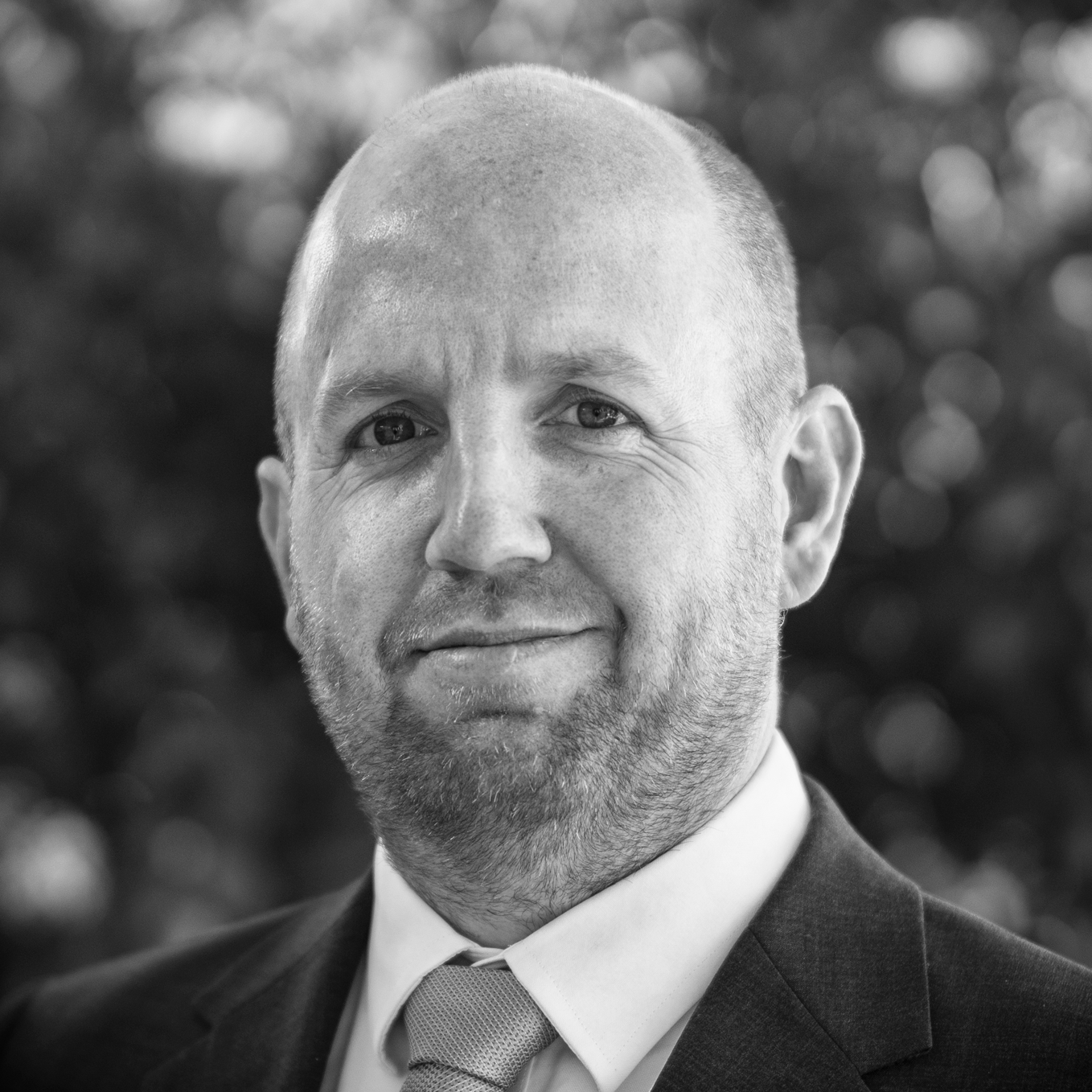 Black and white close-up portrait of a middle-aged man with short hair, wearing a suit and tie, outdoors with blurred foliage in the background.