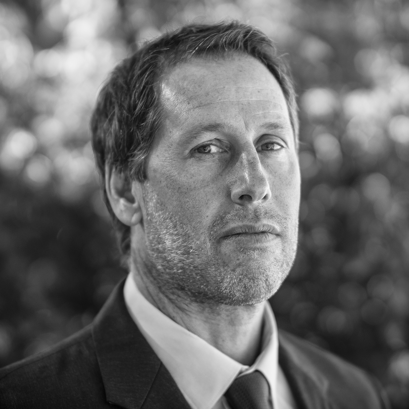 Black-and-white close-up portrait of a man in a suit looking at the camera with a serious expression, background of blurred trees.