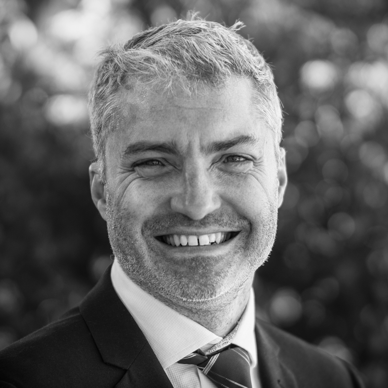 Black and white headshot photograph of a smiling middle-aged man with short, slightly tousled hair, wearing a suit jacket, dress shirt, and tie, against a blurred outdoor background.