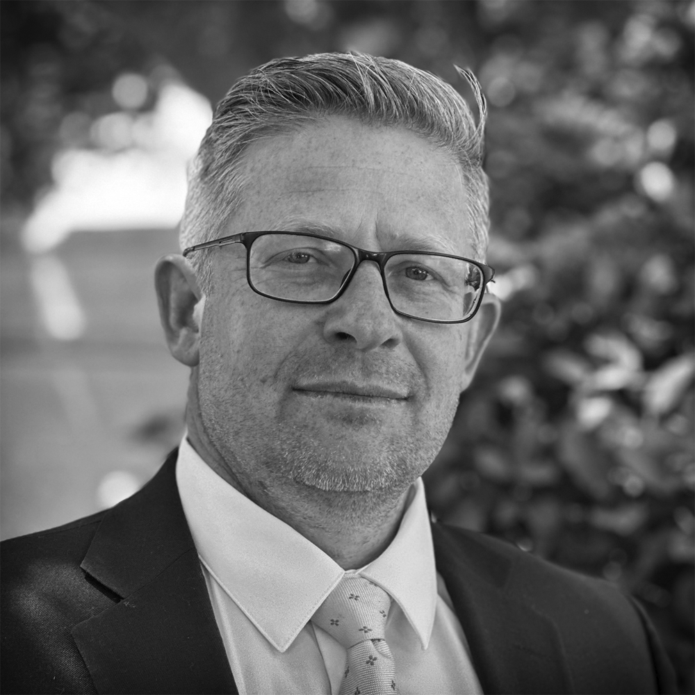 Black and white close-up portrait of a middle-aged man with glasses, short gray hair, wearing a suit and tie, outdoors with blurred foliage in the background.