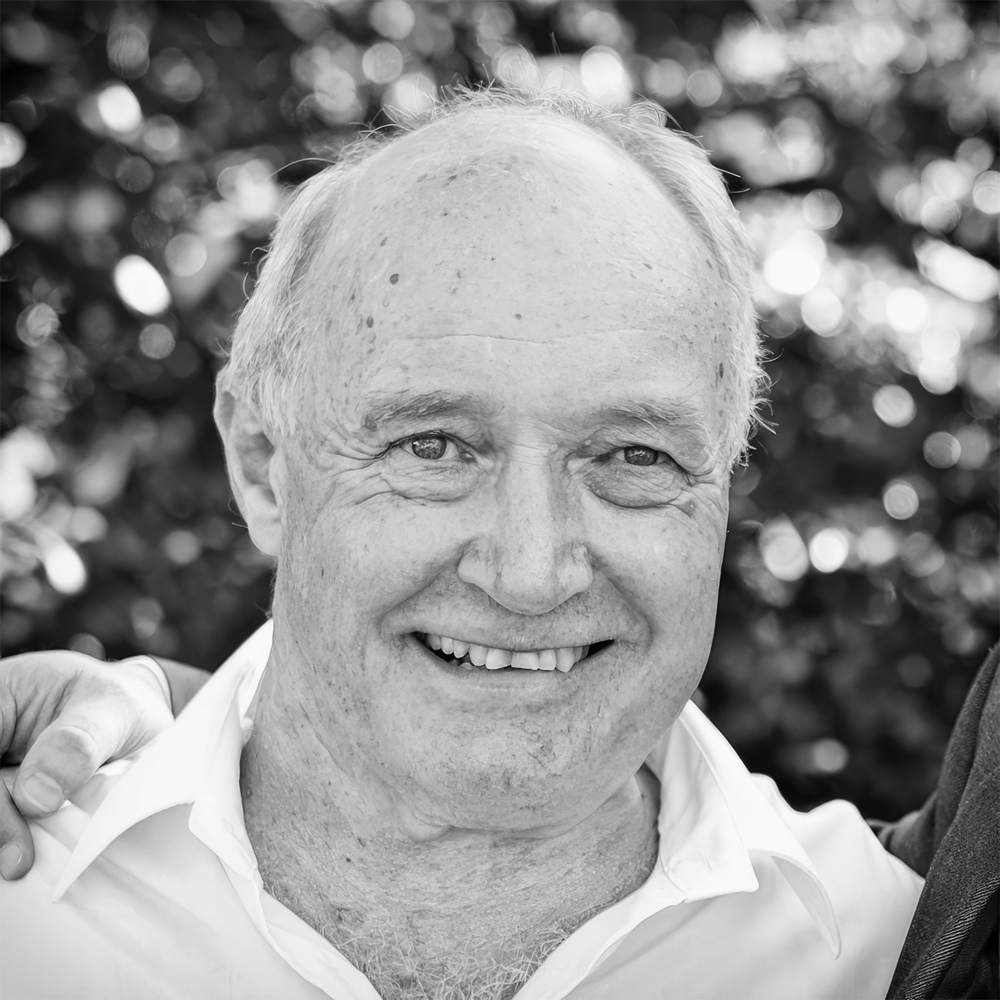 Close-up black-and-white photograph of smiling older man with light hair and white shirt, outdoors with blurred leafy background.
