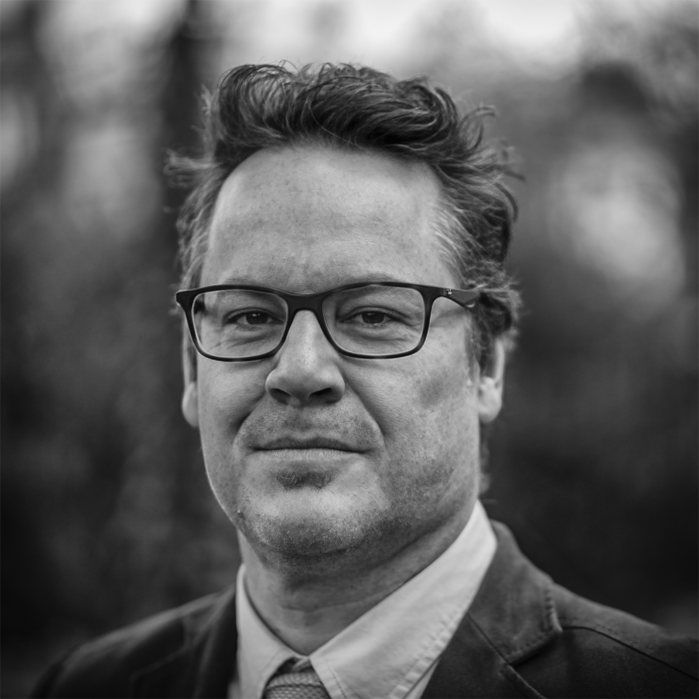 Black and white portrait of a man with glasses, wearing a suit and tie, outdoors with a blurred background.