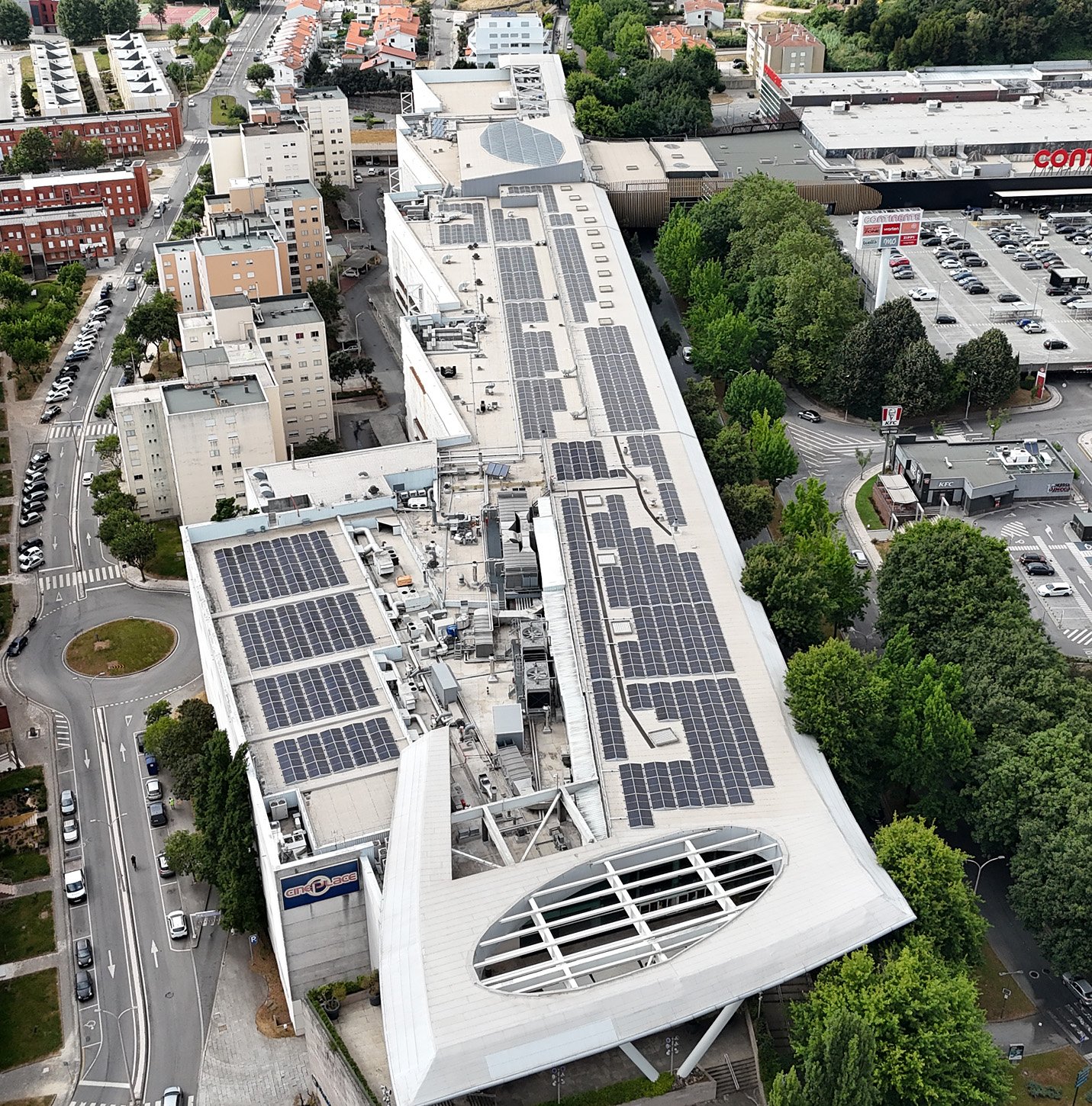 Aerial view of a large building with numerous solar panels on its roof, surrounded by roads, parking lots, trees, and nearby buildings.