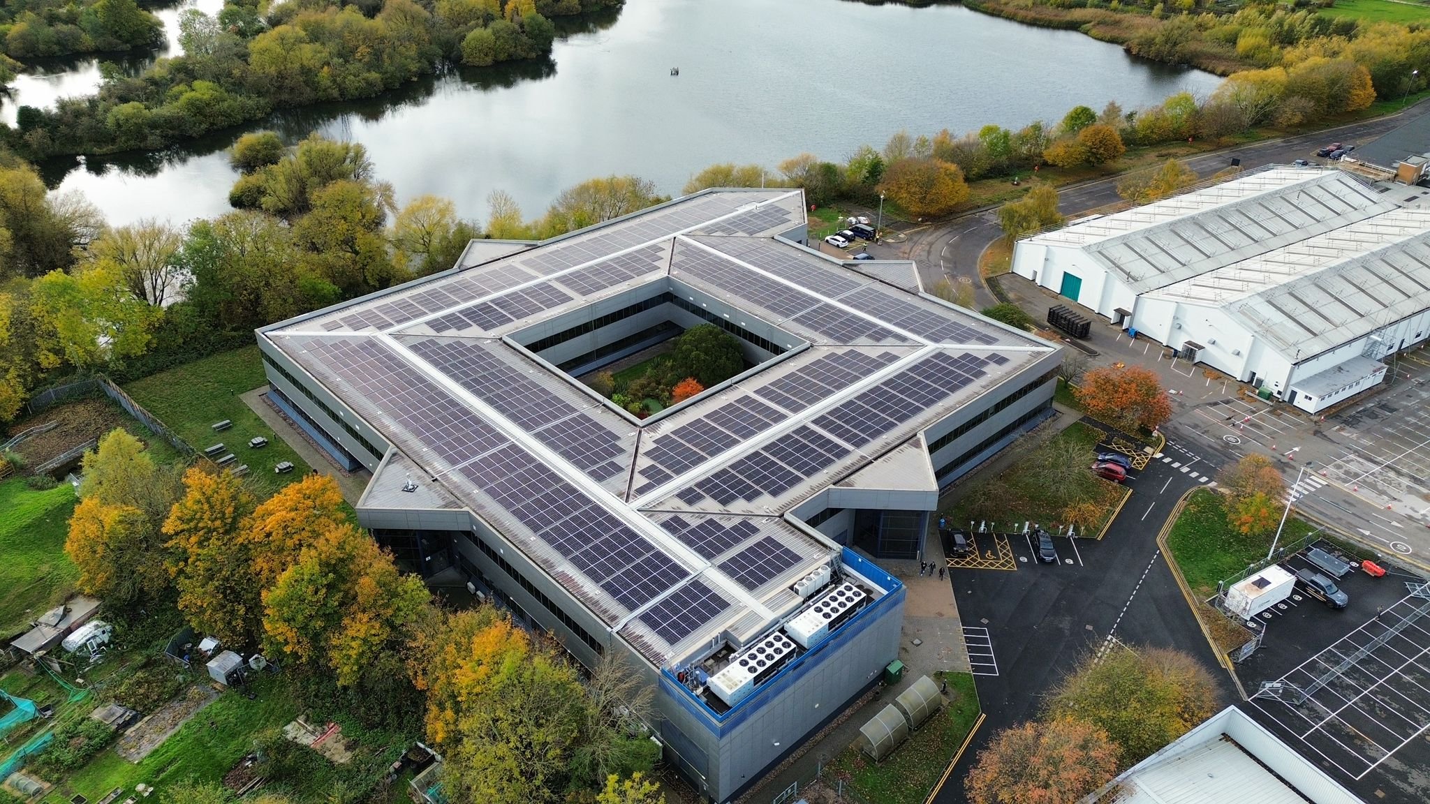 Aerial view of a modern office building with a roof covered in solar panels, surrounded by trees, a pond, and additional industrial buildings in the background.