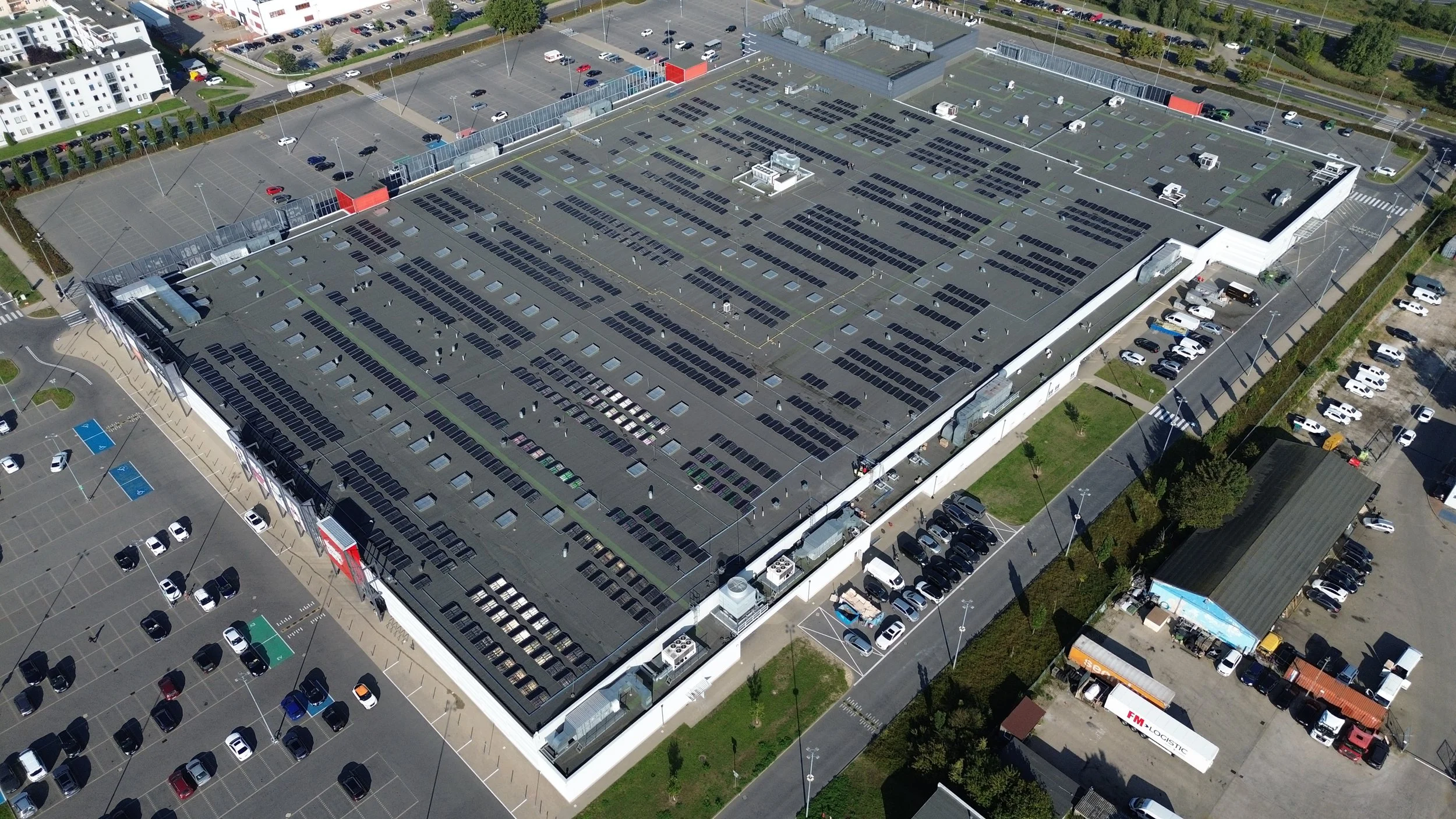 Aerial view of a large retail store with a rooftop parking lot filled with numerous parked cars. Surrounding the store are additional parking areas, some with marked handicapped spots, and various surrounding buildings, trees, and roads.
