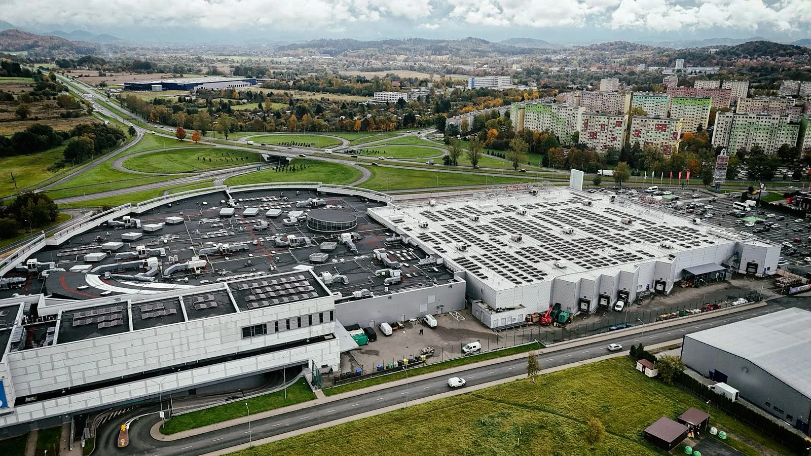 Aerial view of a large commercial building with a parking lot and surrounding roads, with a cityscape and mountains in the background.