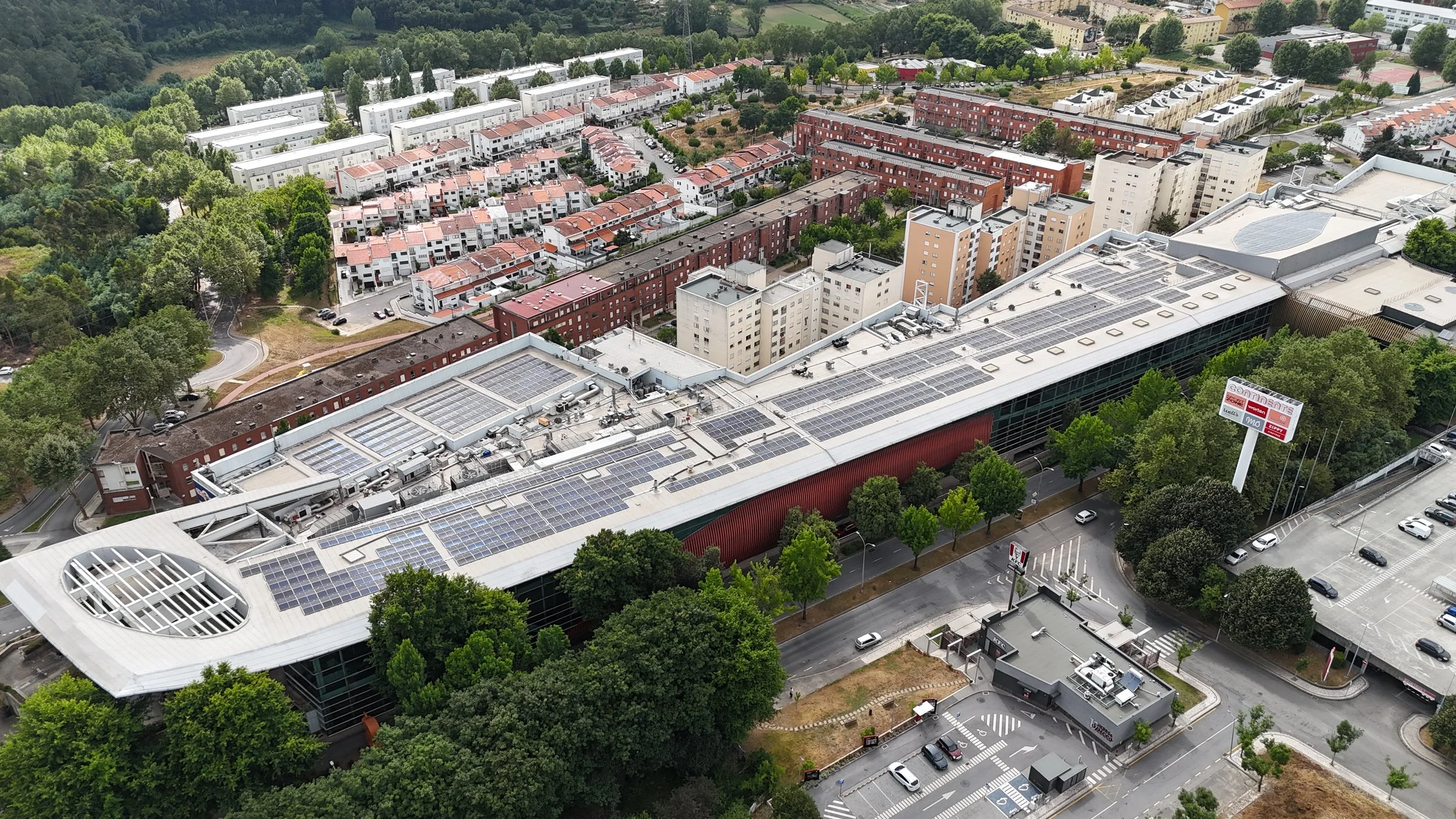 Aerial view of a commercial building with solar panels on the roof, surrounded by trees, parking lot, and residential areas with red and white buildings.