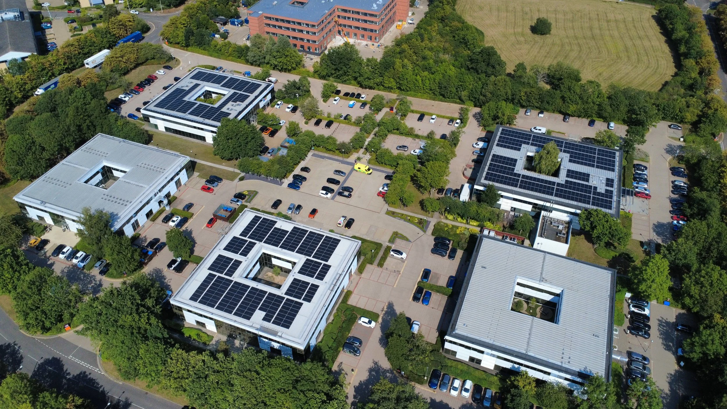 Aerial view of multiple office buildings with solar panels on roofs, surrounded by parking lots, trees, and nearby green spaces.
