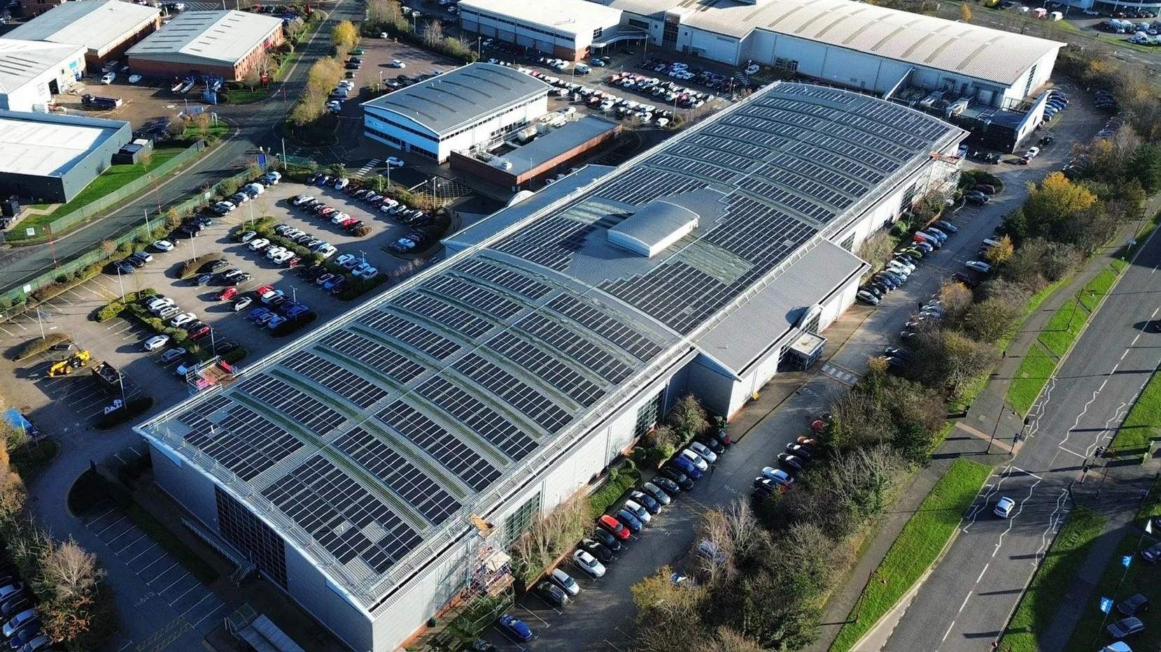 An aerial view of a commercial complex with large buildings, parking lots filled with cars, and a solar panel-covered roof.