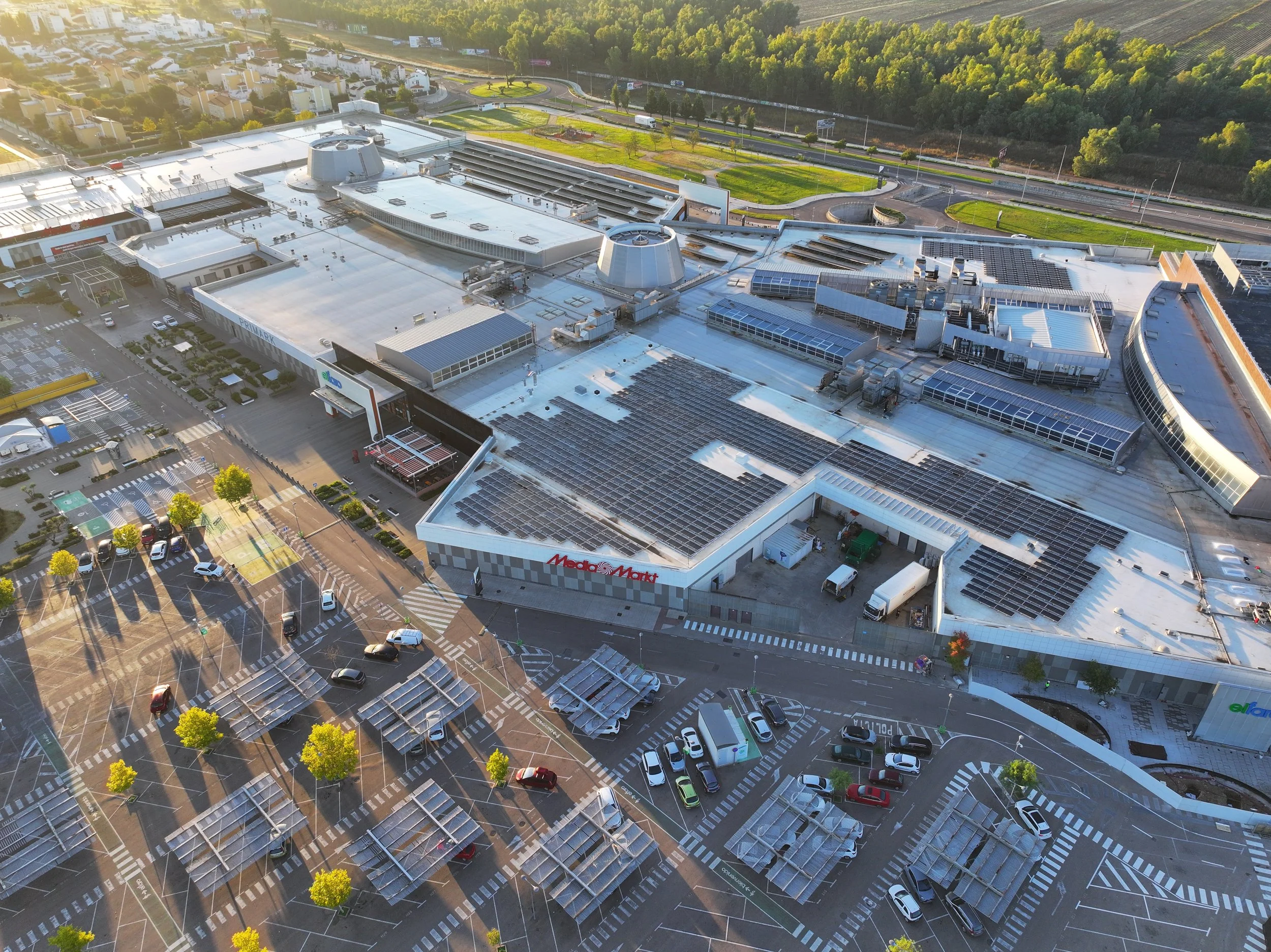 Aerial view of a large shopping mall with parking lots, solar panels on the roofs, and surrounding roads with cars and trees in daylight.