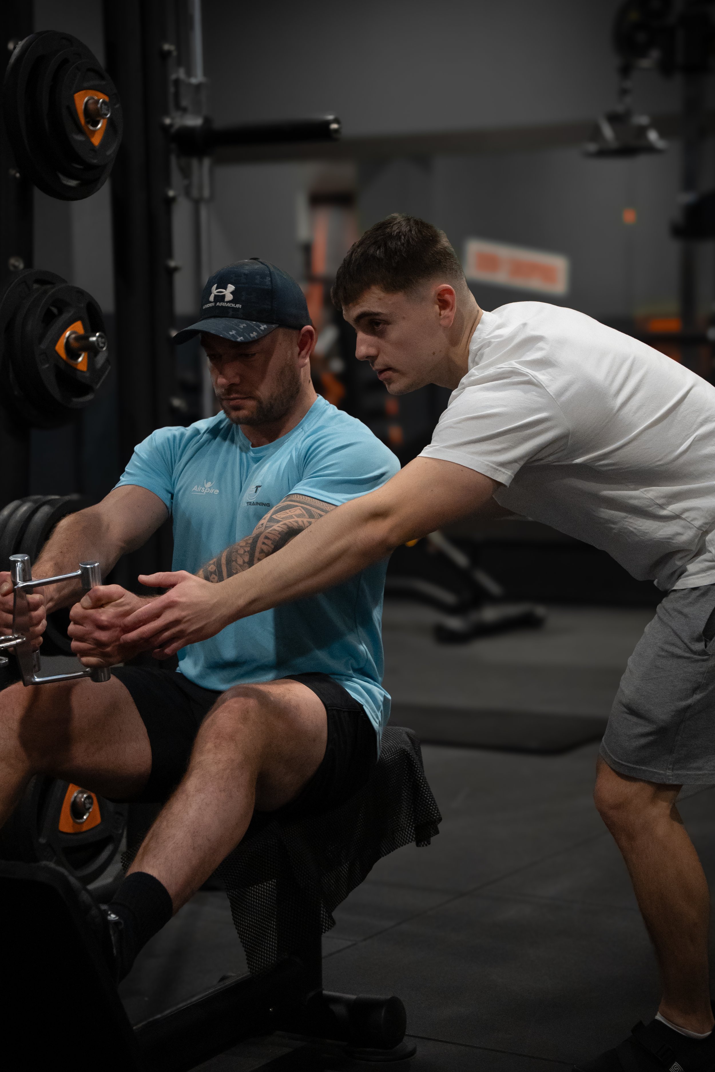 Deux hommes dans une salle de sport. L'un est assis et utilise une machine de musculation, tandis que l'autre l'aide en tenant ses bras. Il y a des poids en arrière-plan.